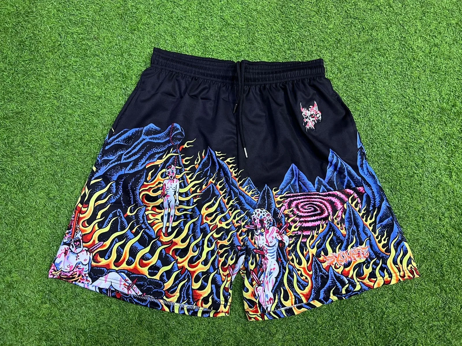 Colorful shorts featuring illustrations of naked women with devil horns surrounded by flames, mountains, and a vortex, on a black background.