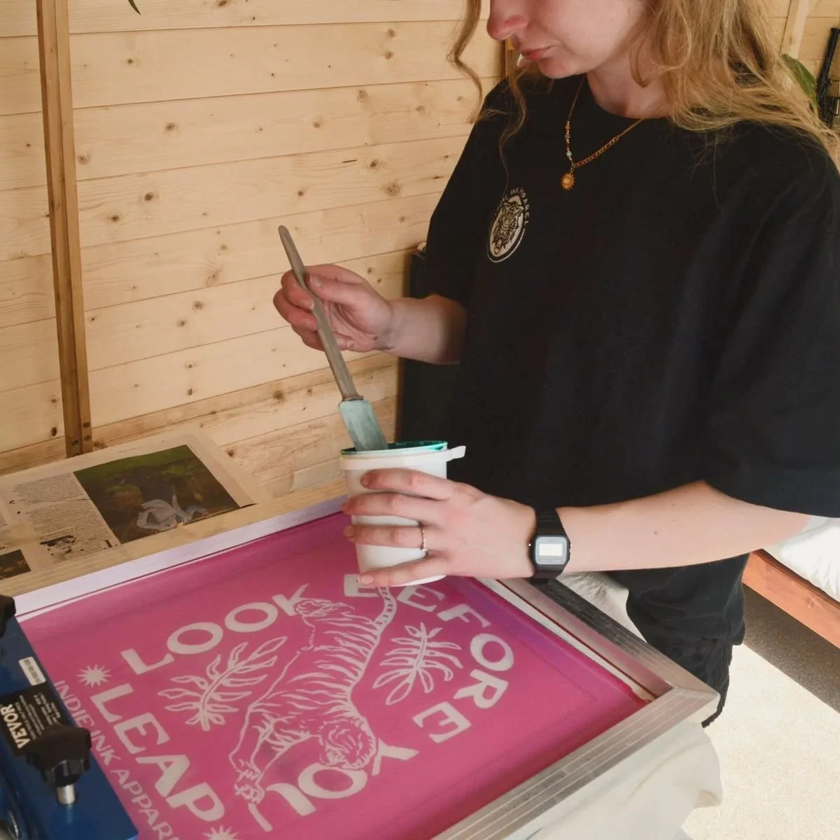 A woman is screen printing a design onto a pink fabric. She is holding a squeegee and a container of ink. The design features a tiger and the words "One Look" and "You." She is in a room with wooden walls, and magazines are on the table next to her.