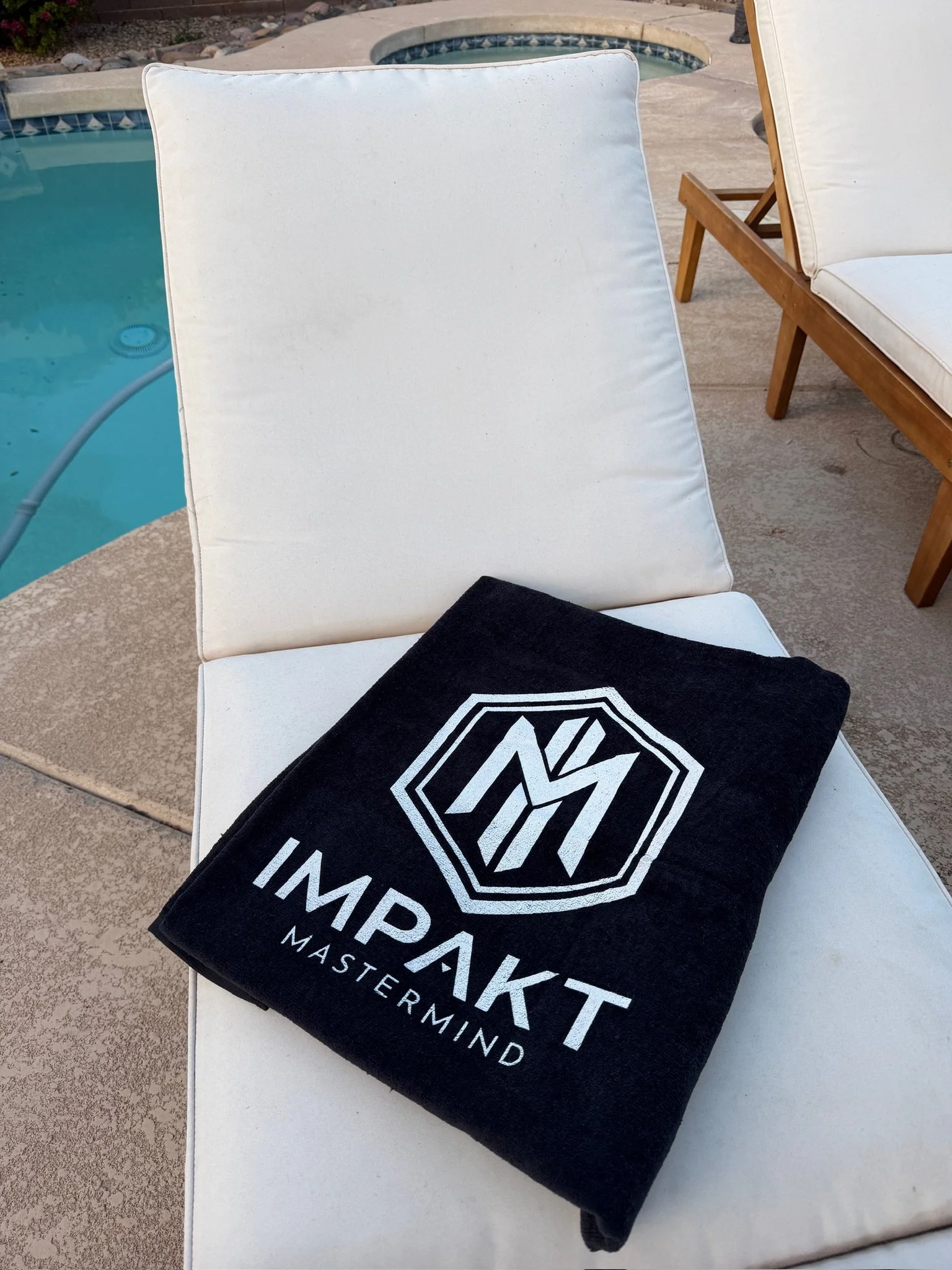 A white lounge chair by a swimming pool with a black towel that has a white logo and the words 'IMPAKT MASTER MIND' on it.