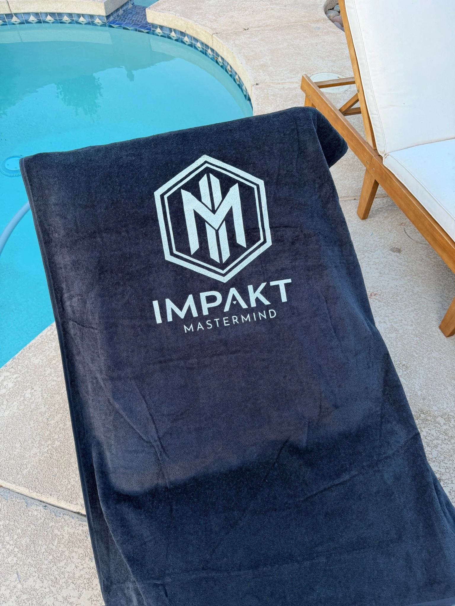 A black towel with the logo and text of Impact Mastermind laid on a poolside lounge chair. The background shows a swimming pool and another lounge chair with a white cushion.