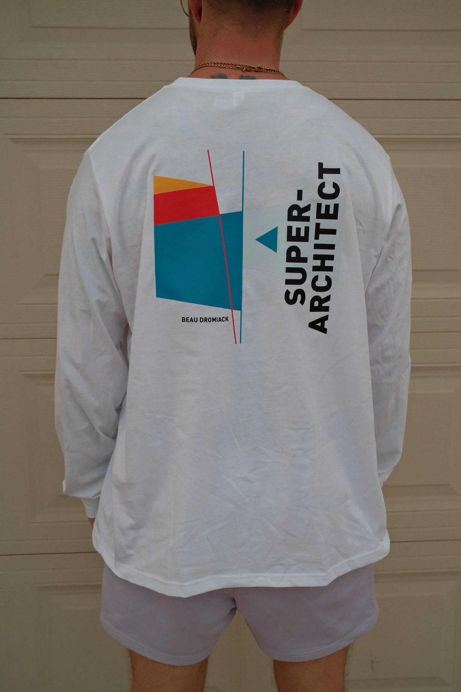 Back of a person wearing a white long-sleeve shirt with a colorful graphic design and text, standing in front of a beige wall.