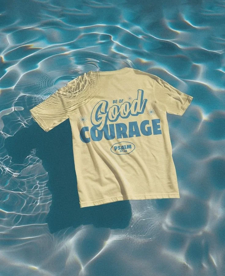 A beige T-shirt with blue text that says 'Be of Good Courage' and includes a reference to Psalm 27:14, floating on the water in a pool.