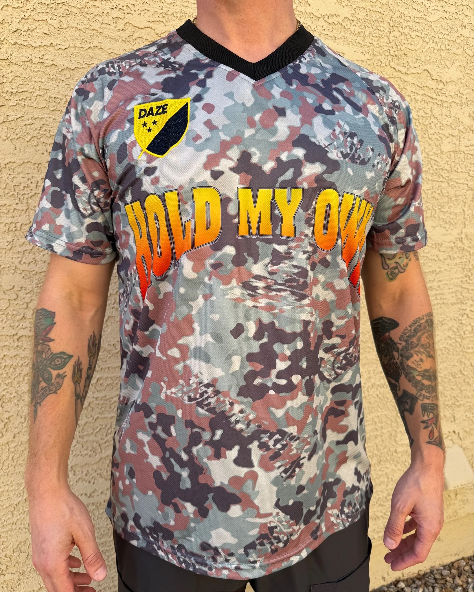 A person wearing a camo patterned shirt with the phrase 'HOLD MY OWN' in large yellow and orange letters on the front. The shirt has patches, one of which says 'DAZE' with a four-star symbol. The person has tattoos on both arms and is standing agains