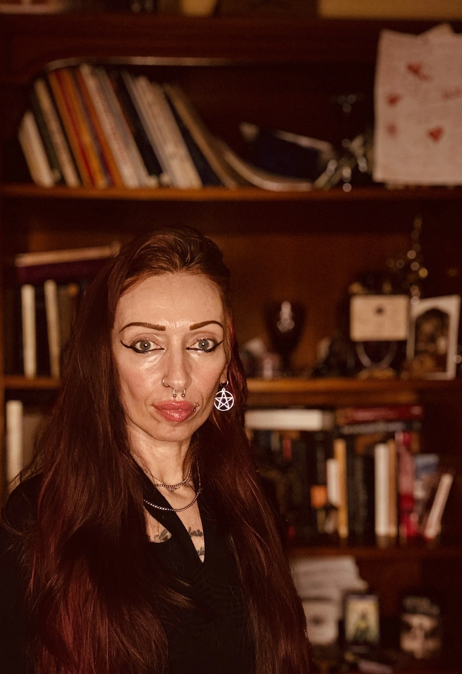 Ragazza con capelli lunghi e rossi, trucco marcato, cerchietti con pentacolo, in una stanza con scaffali di libri e oggetti sullo sfondo.