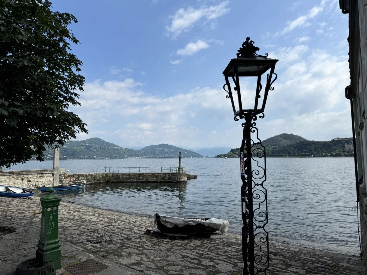Scenic lakeside view with a stone pathway, a black wrought-iron street lamp, a small boat on the shore, a green fire hydrant, and distant hills under a partly cloudy sky in Italy.