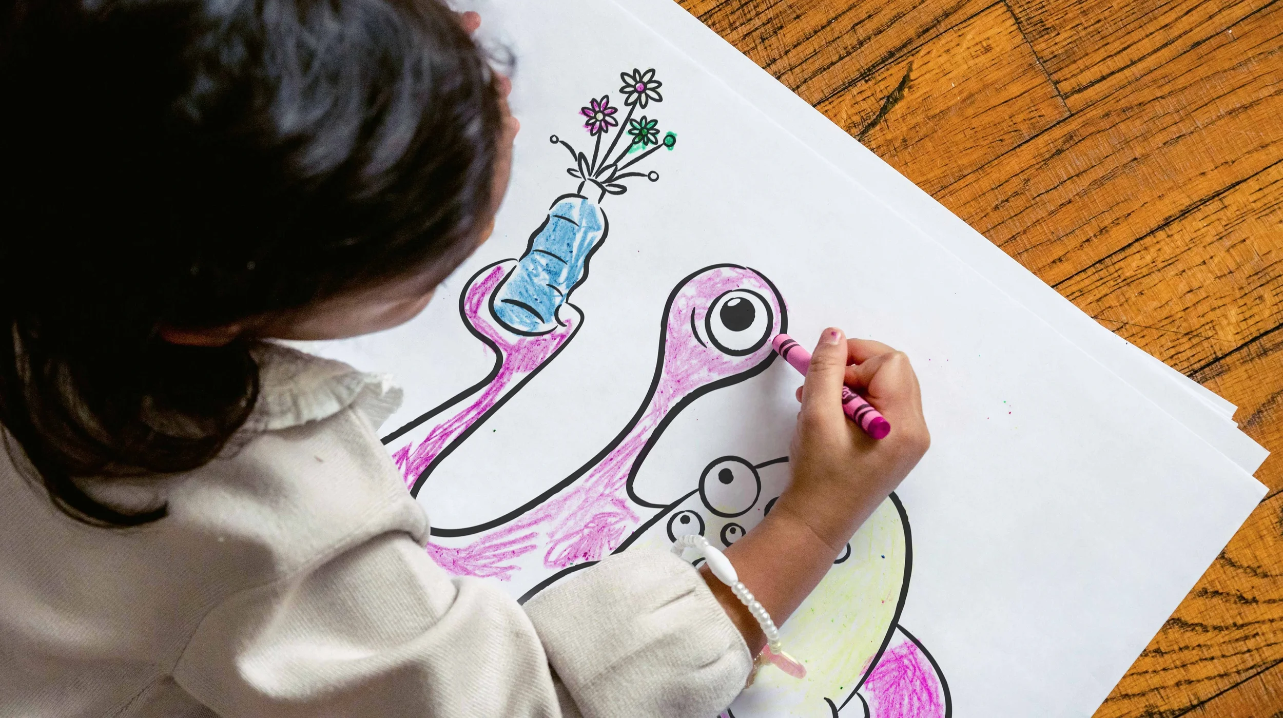 A child coloring a dinosaur drawing with a pink crayon, holding a blue crayon-tipped arm with flowers in a vase on the dinosaur's head.