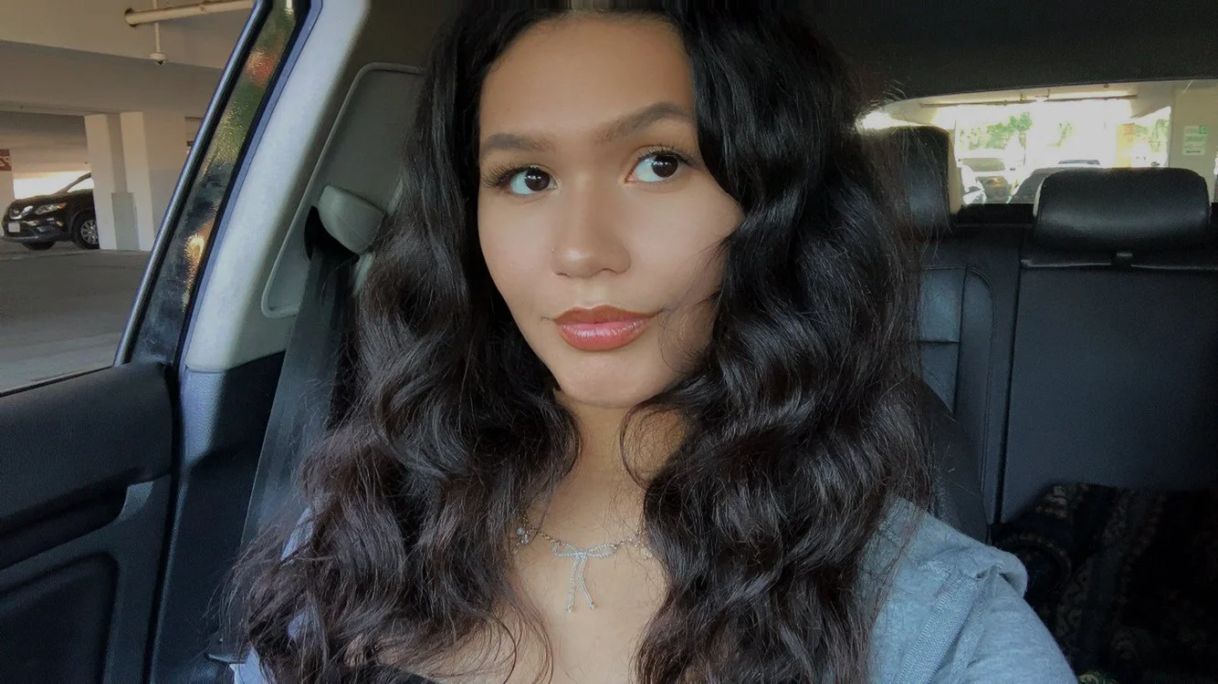 A woman with long, dark hair sits in the back seat of a car, looking thoughtfully out the window