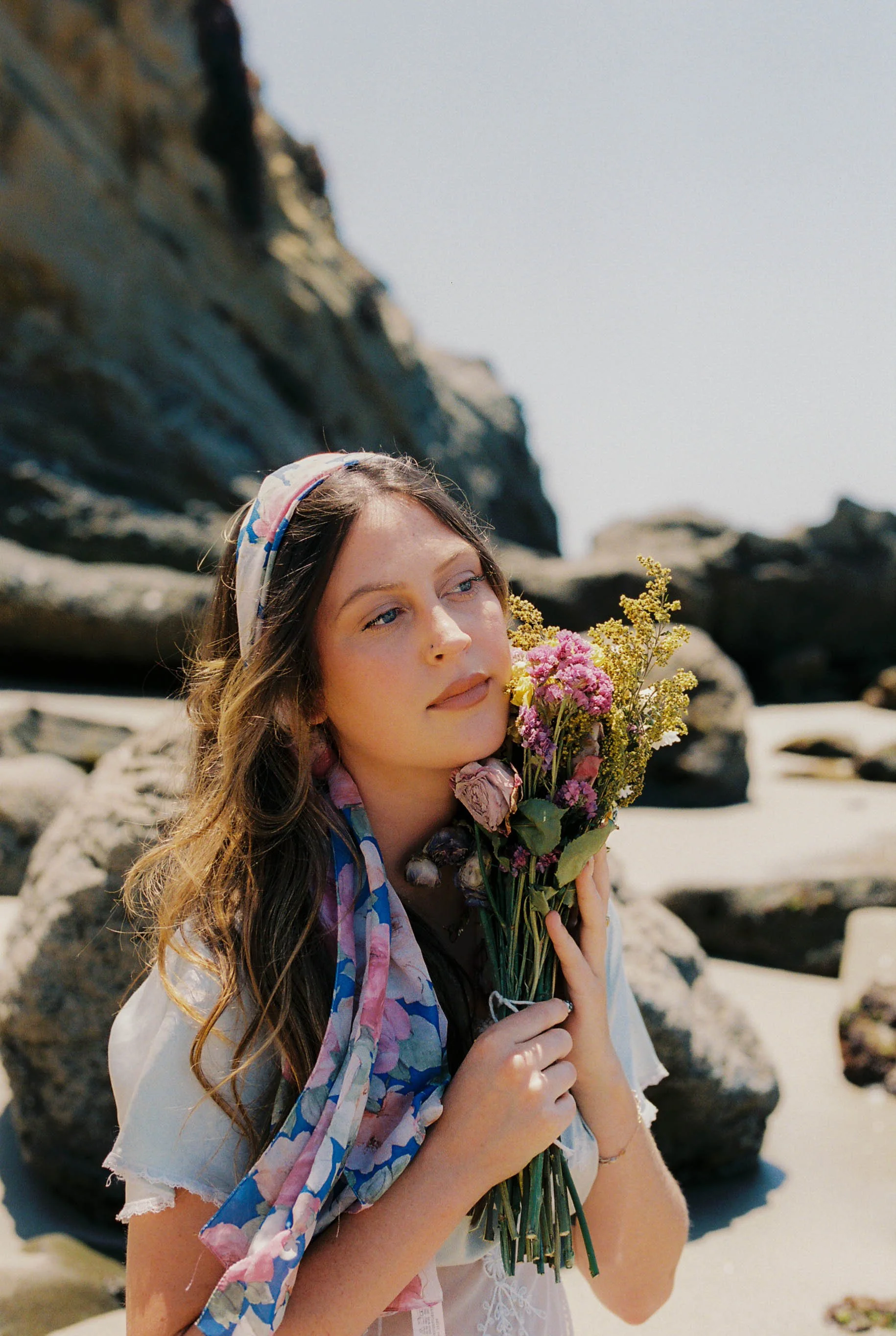 A woman wearing a scarf holds a bouquet of colorful flowers, smiling gently at the camera.
