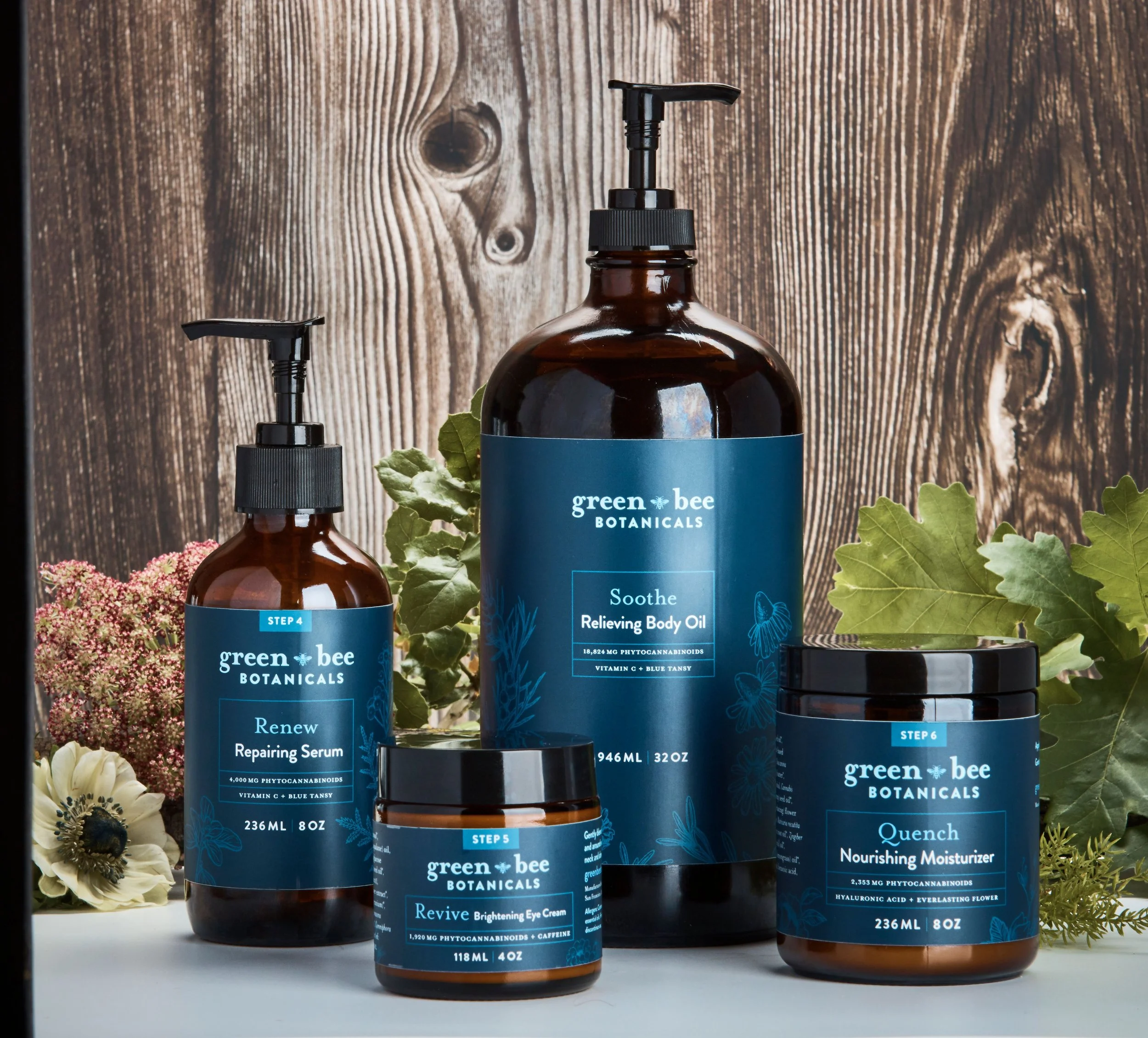 Set of Green Bee Botanicals skincare products including Relief Body Oil, Renew Repairing Serum, Quench Nourishing Moisturizer, Revive Brightening Eye Cream, with flowers and leaves in front of a wooden background.