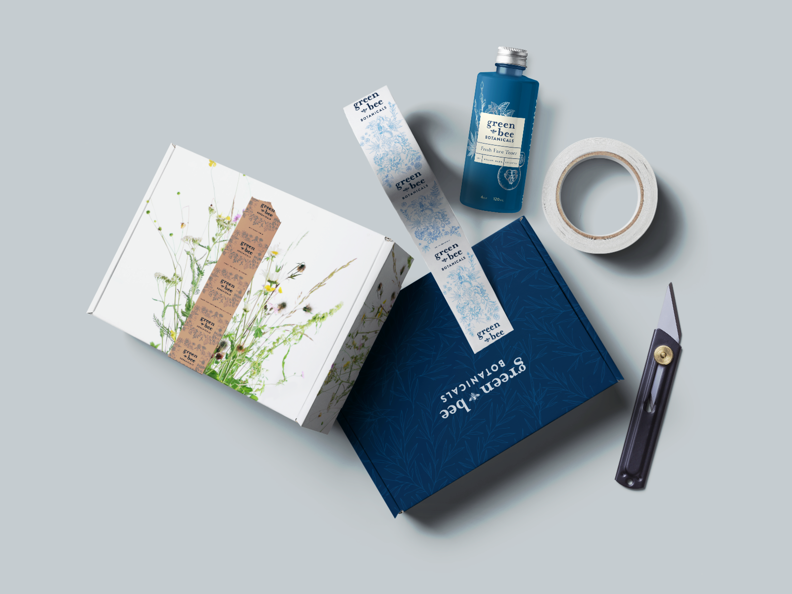 Natural skincare product packaging and tools on a light blue background, including a white box with floral illustrations, a blue box labeled Green Bee Botanicals, a tube of face toner, a roll of masking tape, a box cutter, and a strip of botanical-th