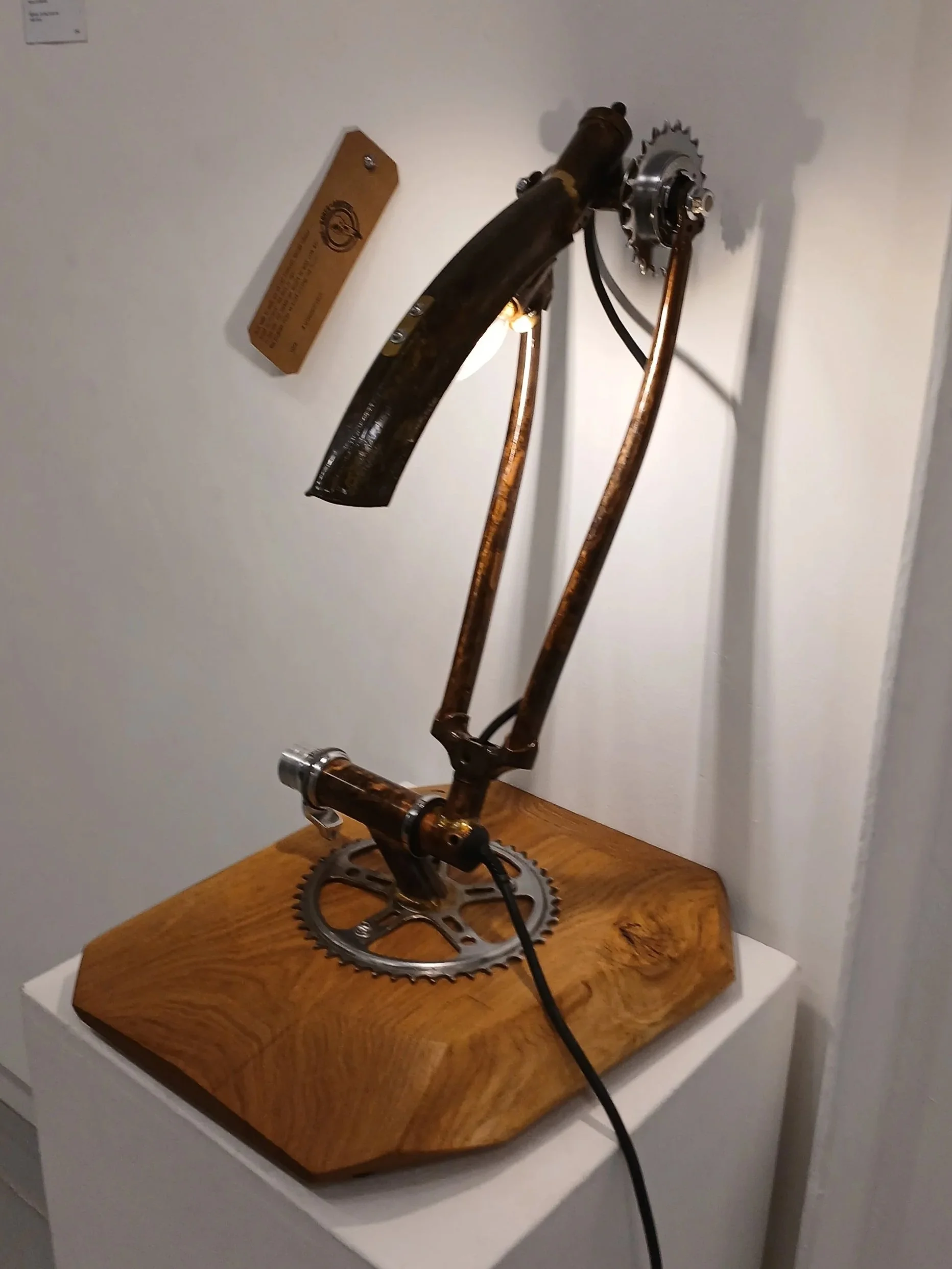 Bike component sculpture mounted on a wooden base, made from bike parts including a chainring, crank arm, and frame, displayed indoors.