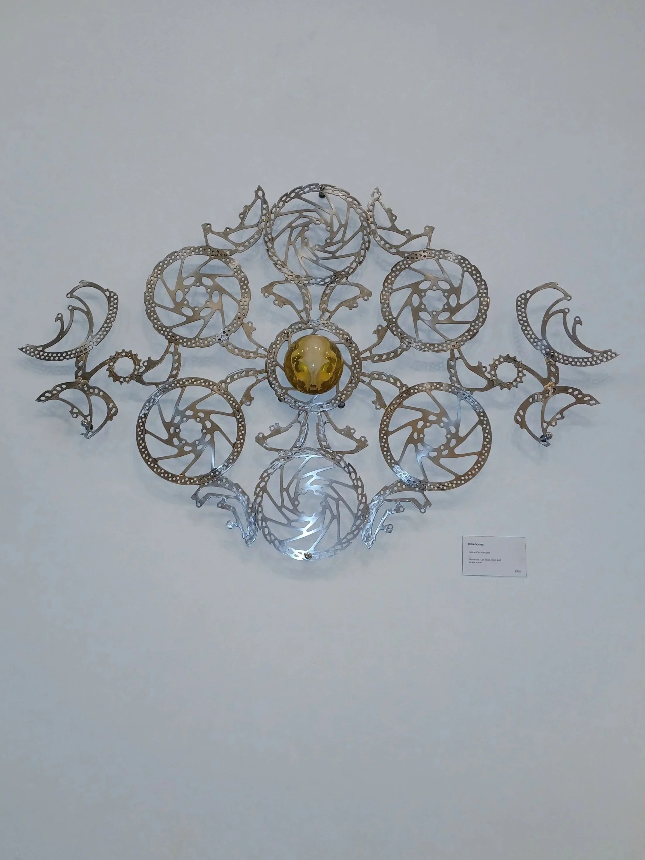 Metal sculpture resembling a four-leaf clover with spiraled circular elements, a yellow and white glass orb at the center, mounted on a white wall in an art gallery.