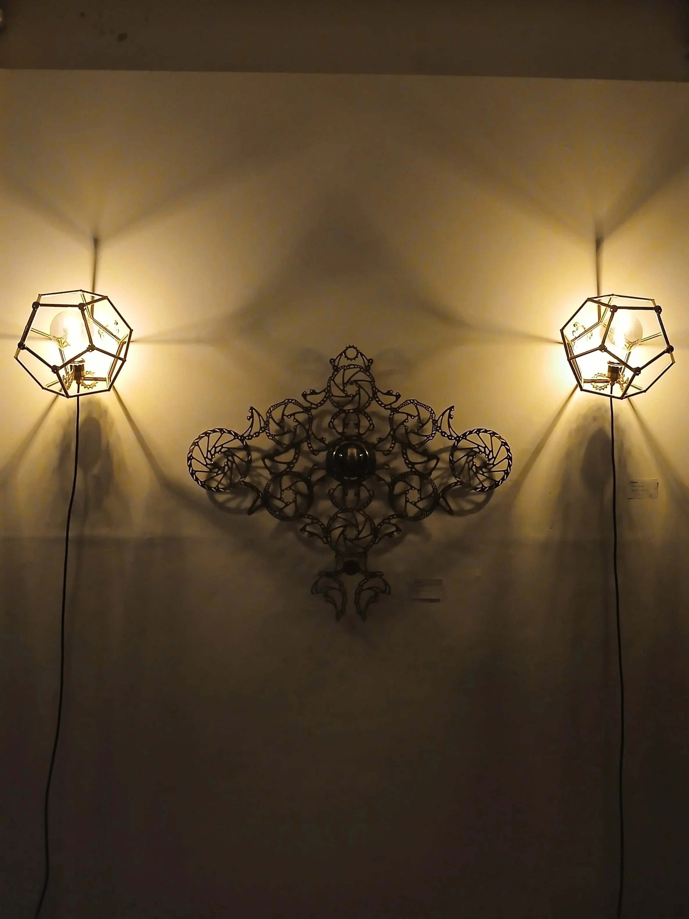 Two wall-mounted light fixtures with geometric metal frames, casting shadows on the wall, with an ornate decorative metal piece in the center.