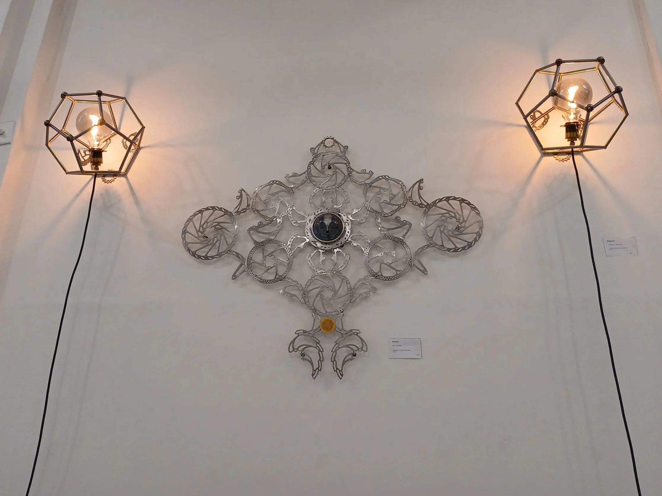 Contemporary wall sculpture with metal rings and scrollwork, flanked by two geometric wall-mounted lamps with exposed bulbs.