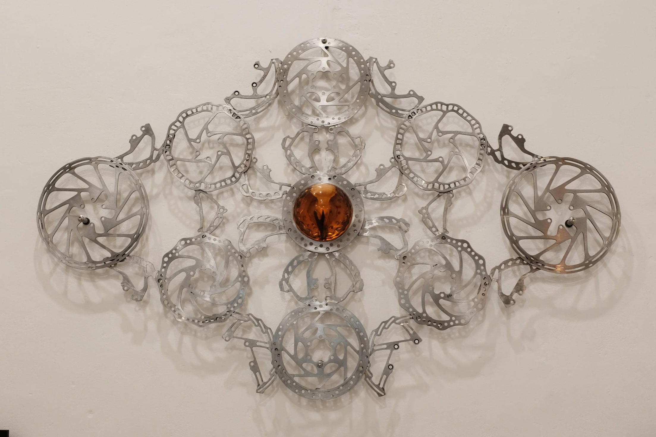 Art installation made of bicycle brake discs and other metal parts arranged in a symmetrical pattern with a central amber-colored sphere with a real crow skull casted in resin