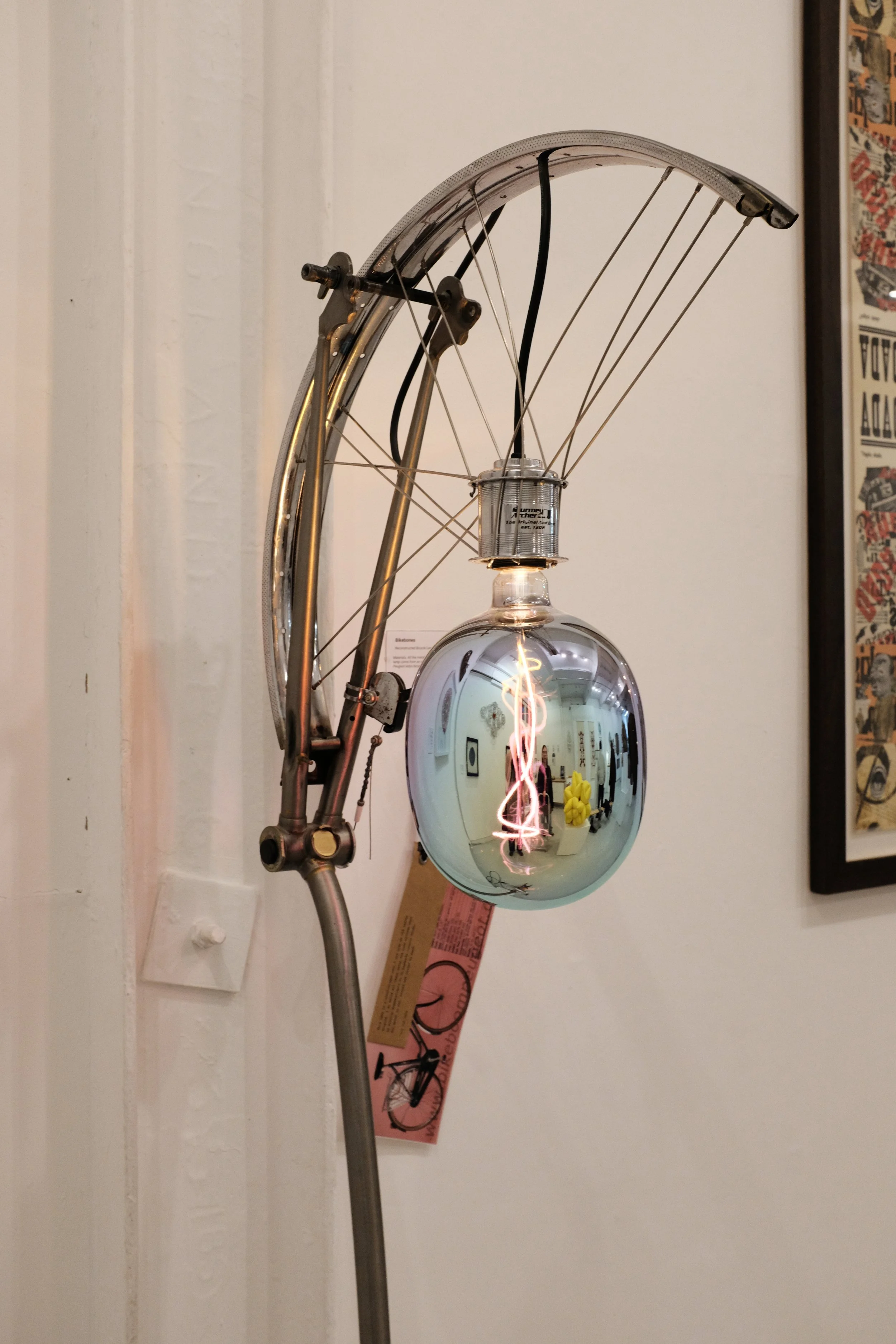 A metallic lamp with a round reflective bulb and an exposed filament, mounted on a wall. The lamp has a curved arm and visible wiring. There is a framed poster or artwork on the wall to the right.