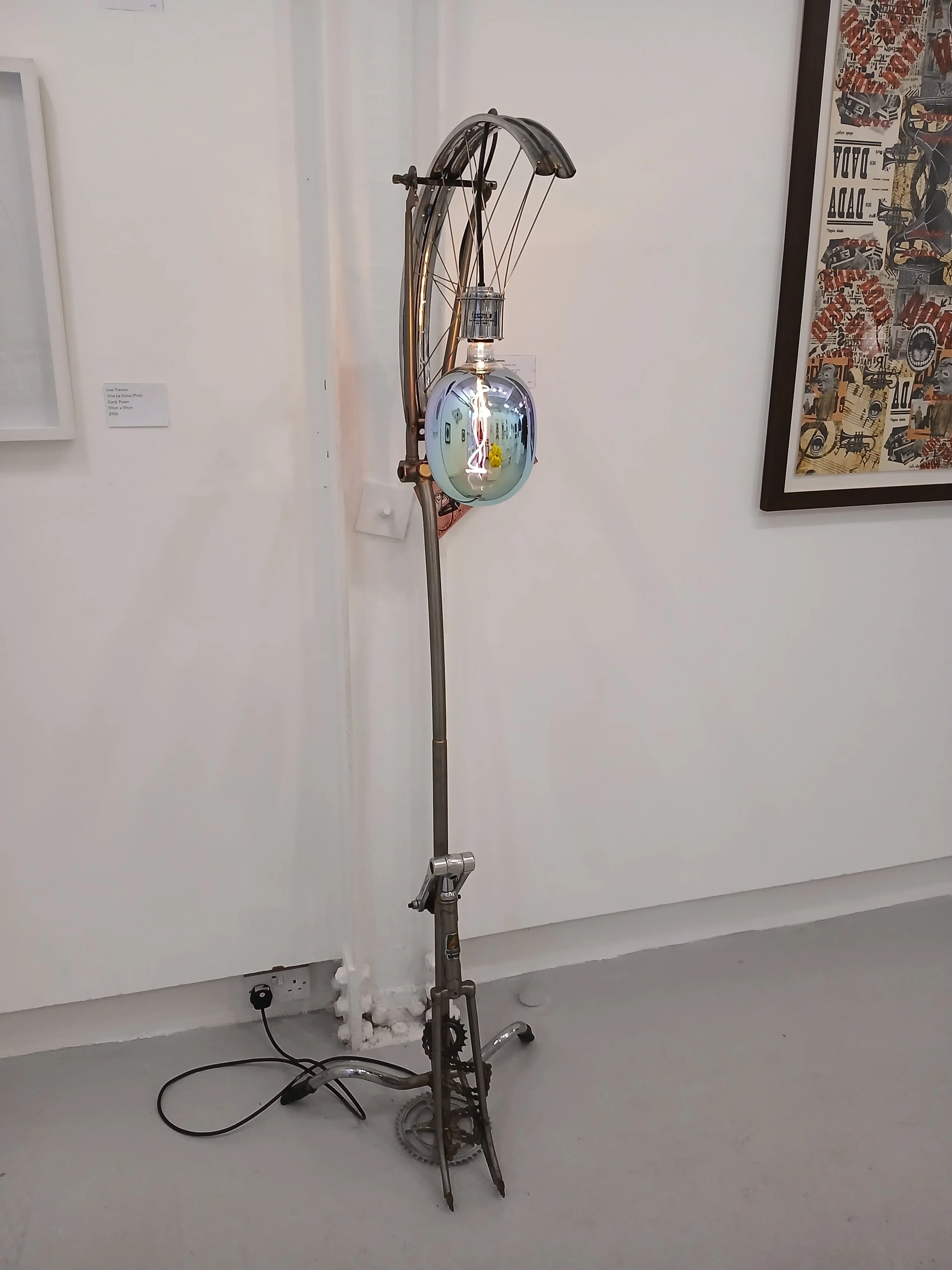 An art installation made from various bicycle parts, including a bike fork, wheel, and frame, arranged with a lamp and reflective sphere on a gray floor in a gallery.