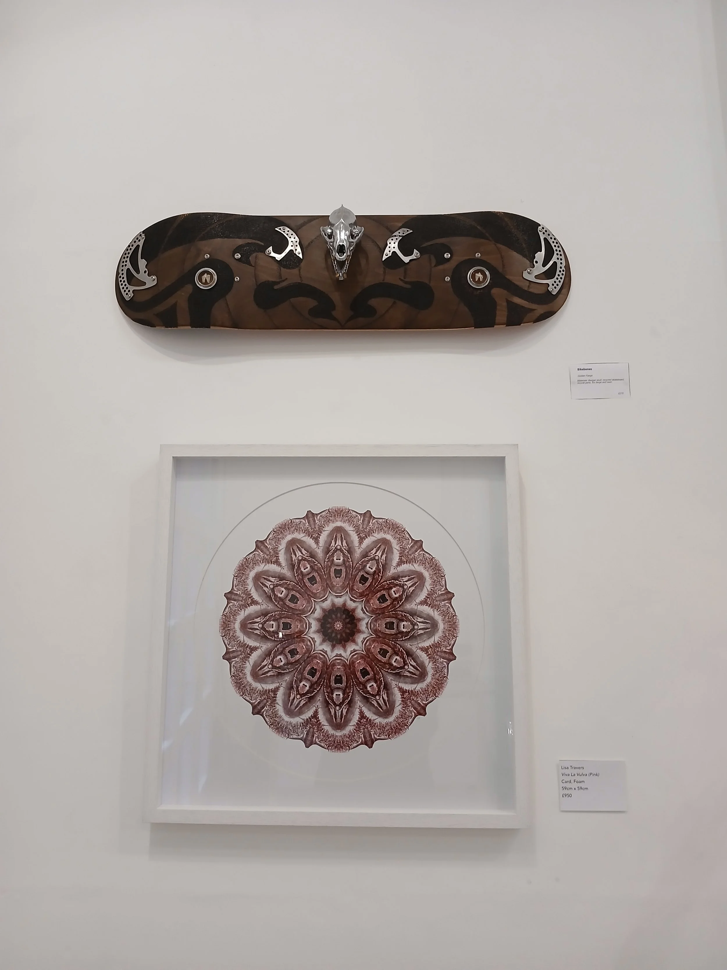 Two pieces of art displayed on a white wall. The upper piece is a skateboard with a dark wood finish and decorative metallic elements, including a skull and crescent shapes. The lower piece is a framed, intricate symmetrical design resembling a pink 