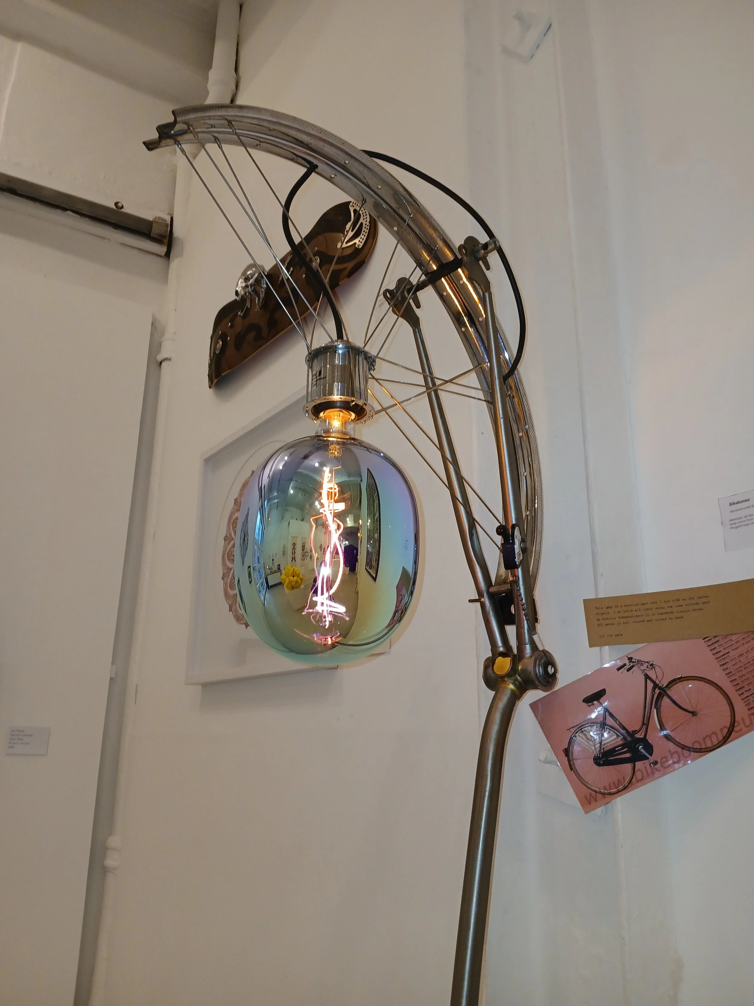 A modern art installation features a bicycle mounted on a wall with an illuminated filament bulb hanging from its frame. The installation includes a decorative element resembling a motorcycle tank, and a reflection of the gallery space can be seen in