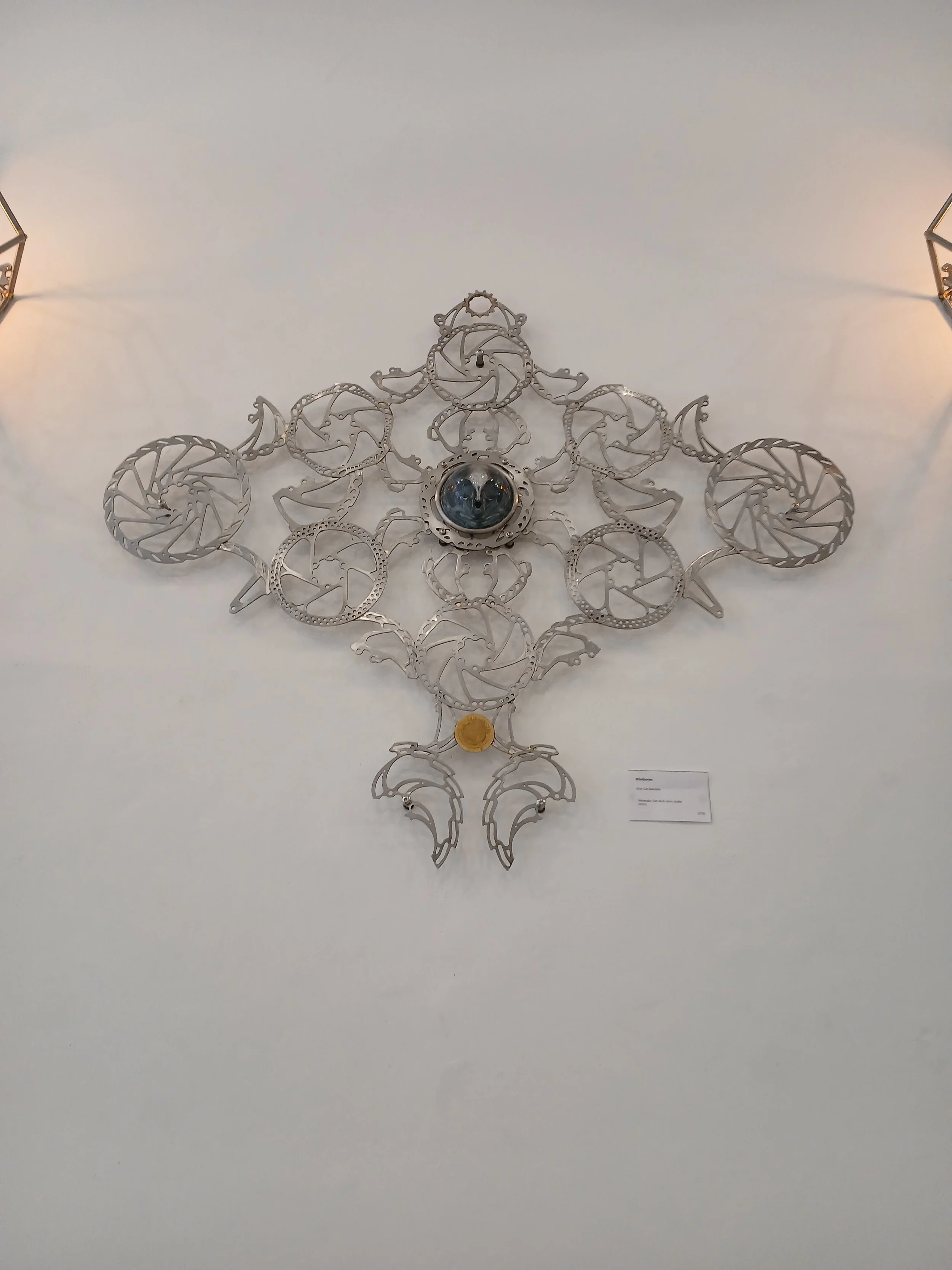 Contemporary artwork made of interconnected silver metallic pieces arranged in an intricate symmetrical design on a white wall. It features a central glass sphere and a small gold coin towards the bottom.