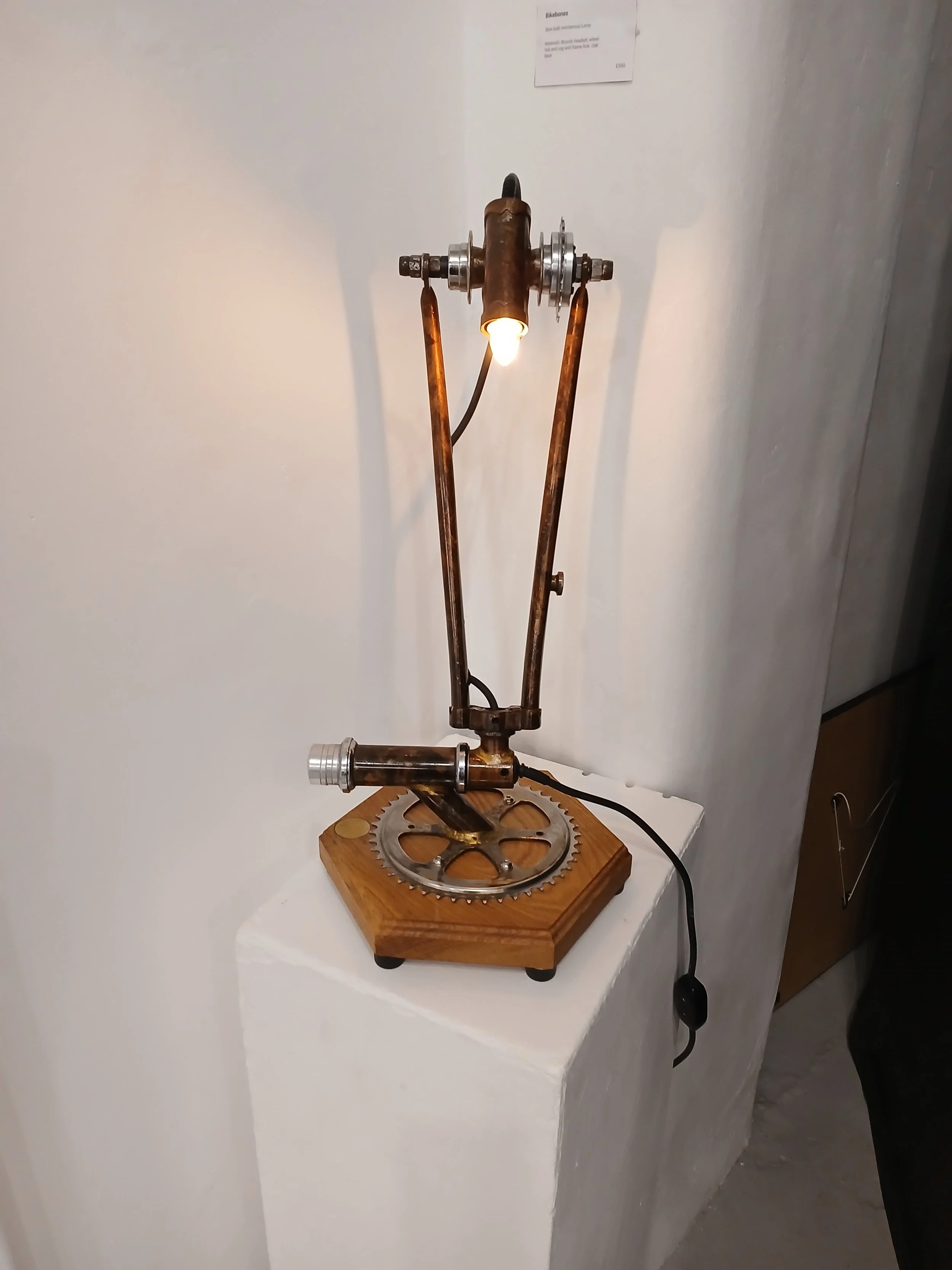 A lamp made from mechanical and metallic parts, including gears, pipes, and a wooden base, displayed on a white pedestal.