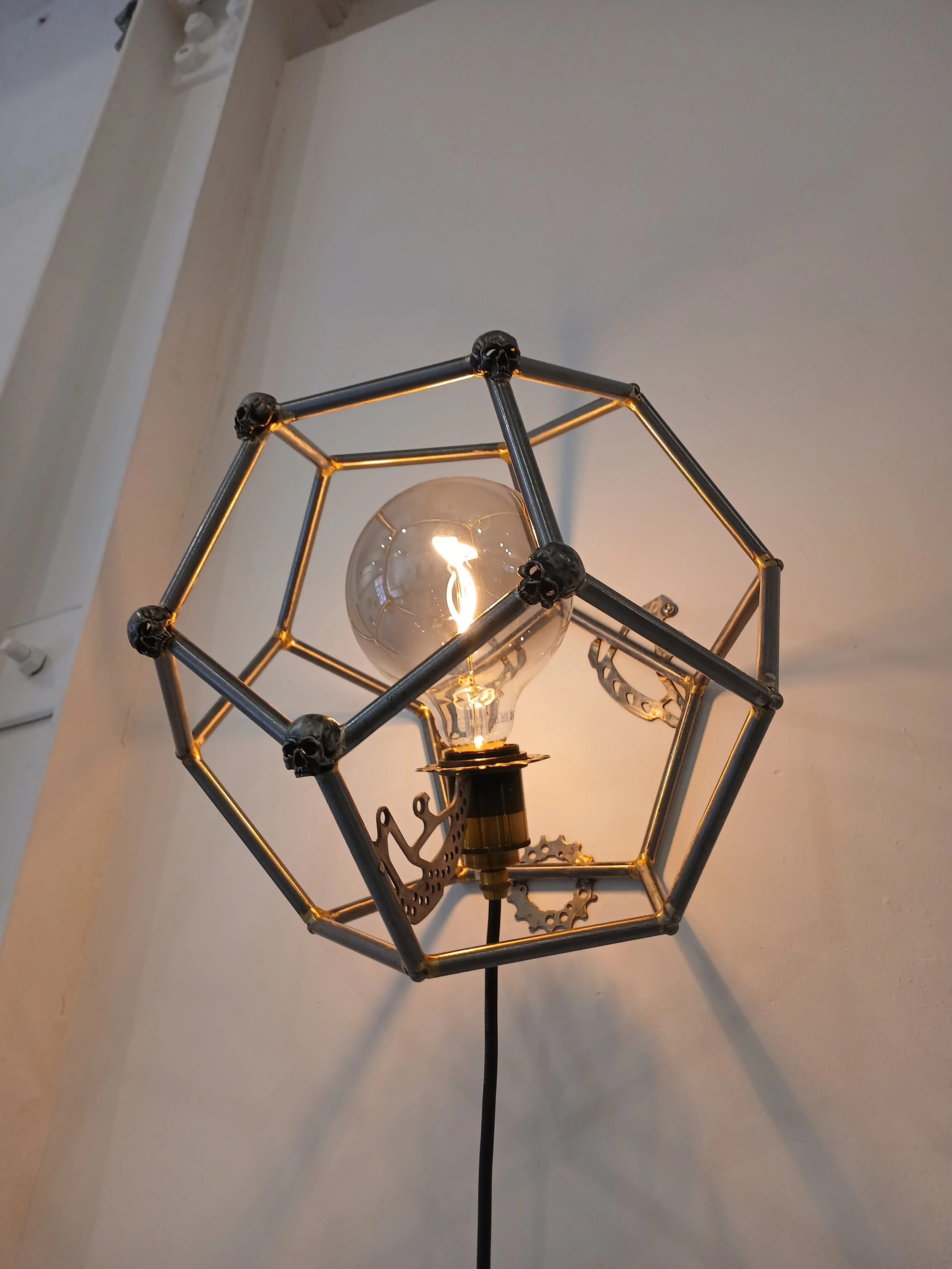 A modern wall-mounted light fixture with an exposed filament bulb inside a geometric metal cage, featuring gear and sprocket decorative elements.