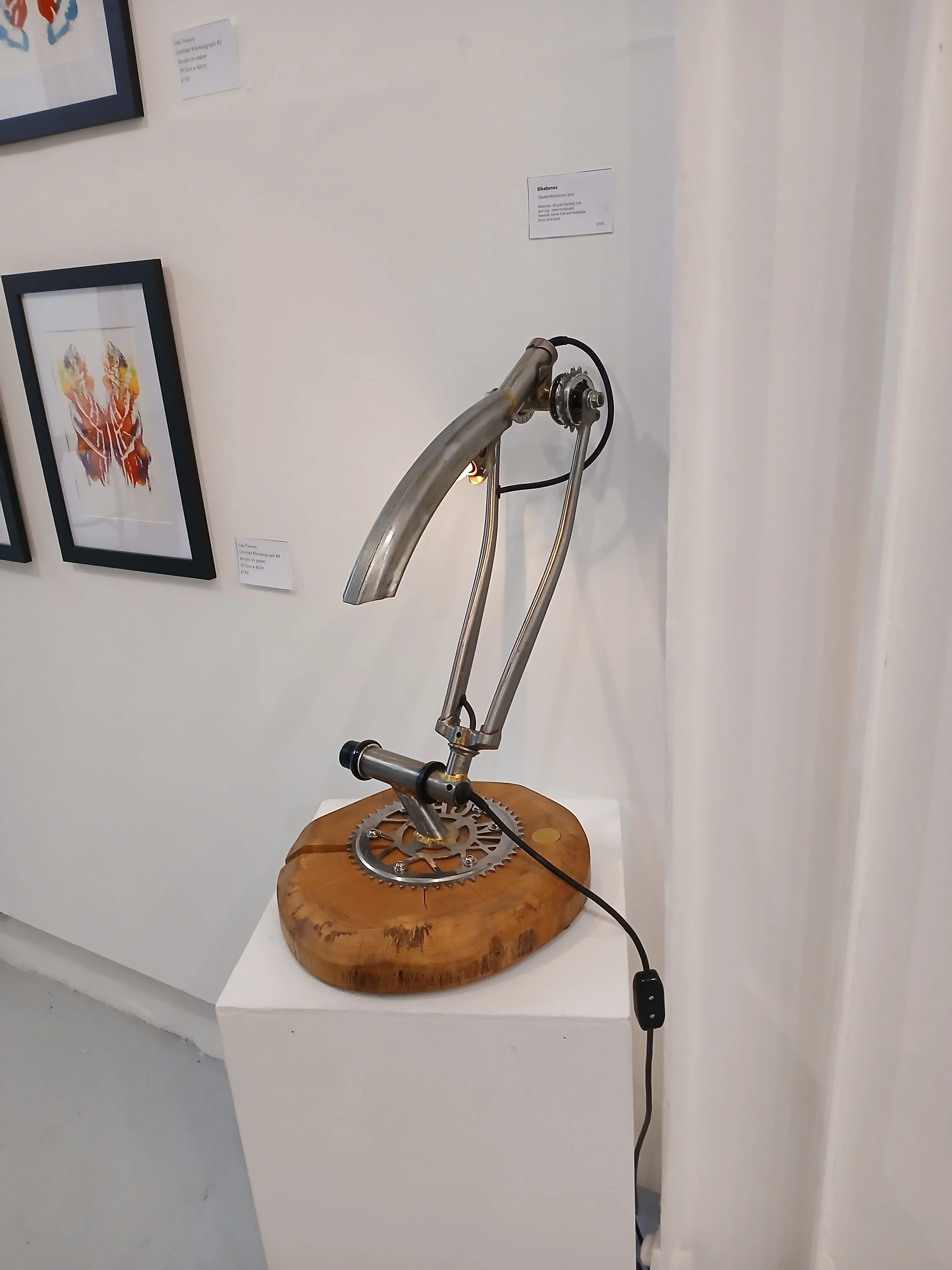 A sculpture made of metal parts including a gear, mounted on a wooden base, displayed on a white pedestal in an art gallery.