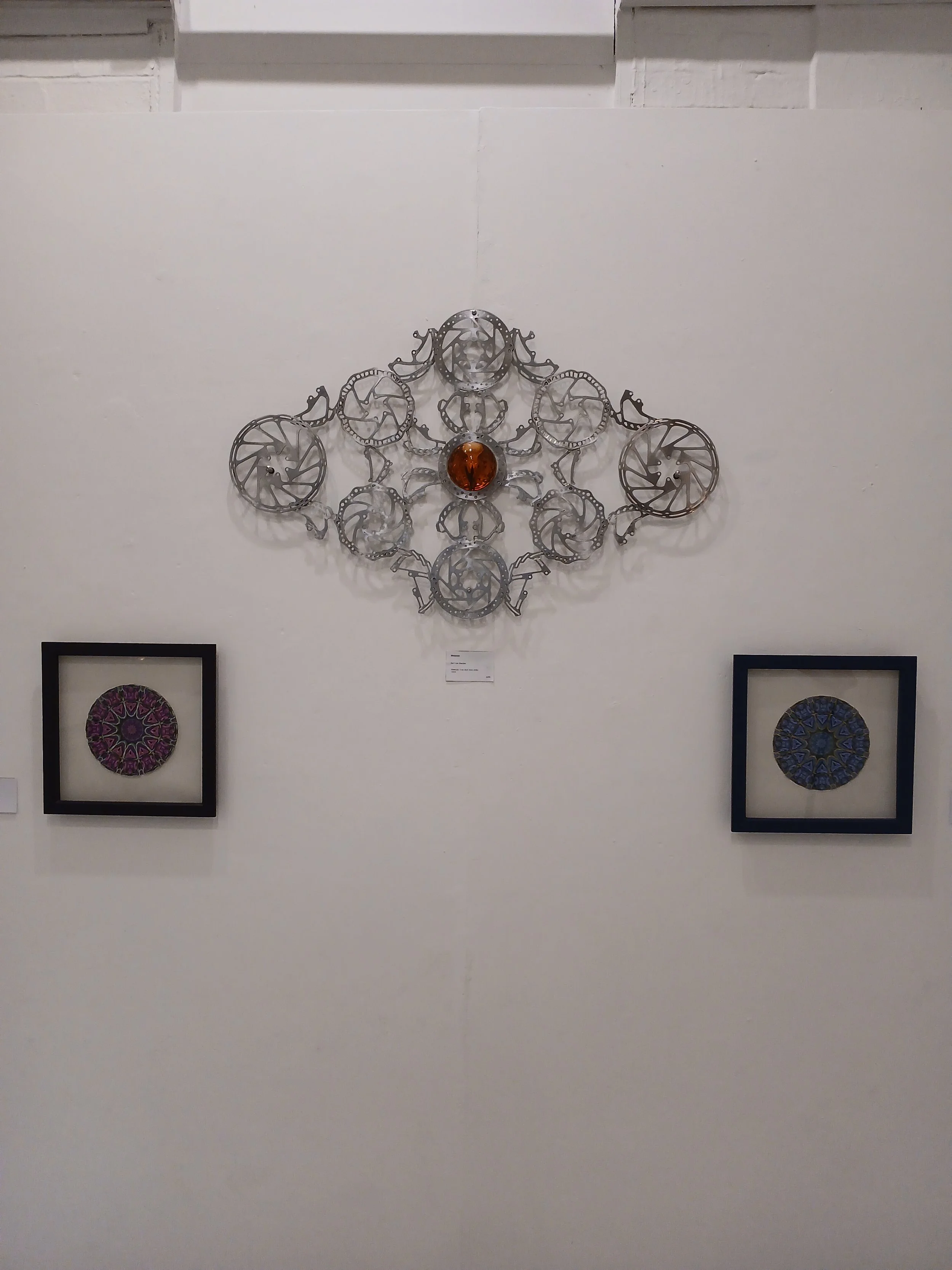 Wall display of modern art with a large metal sculpture in the center, featuring interconnected circular metal pieces with a central orange glass orb, flanked by two framed circular artwork pieces with intricate geometric patterns on the lower left a