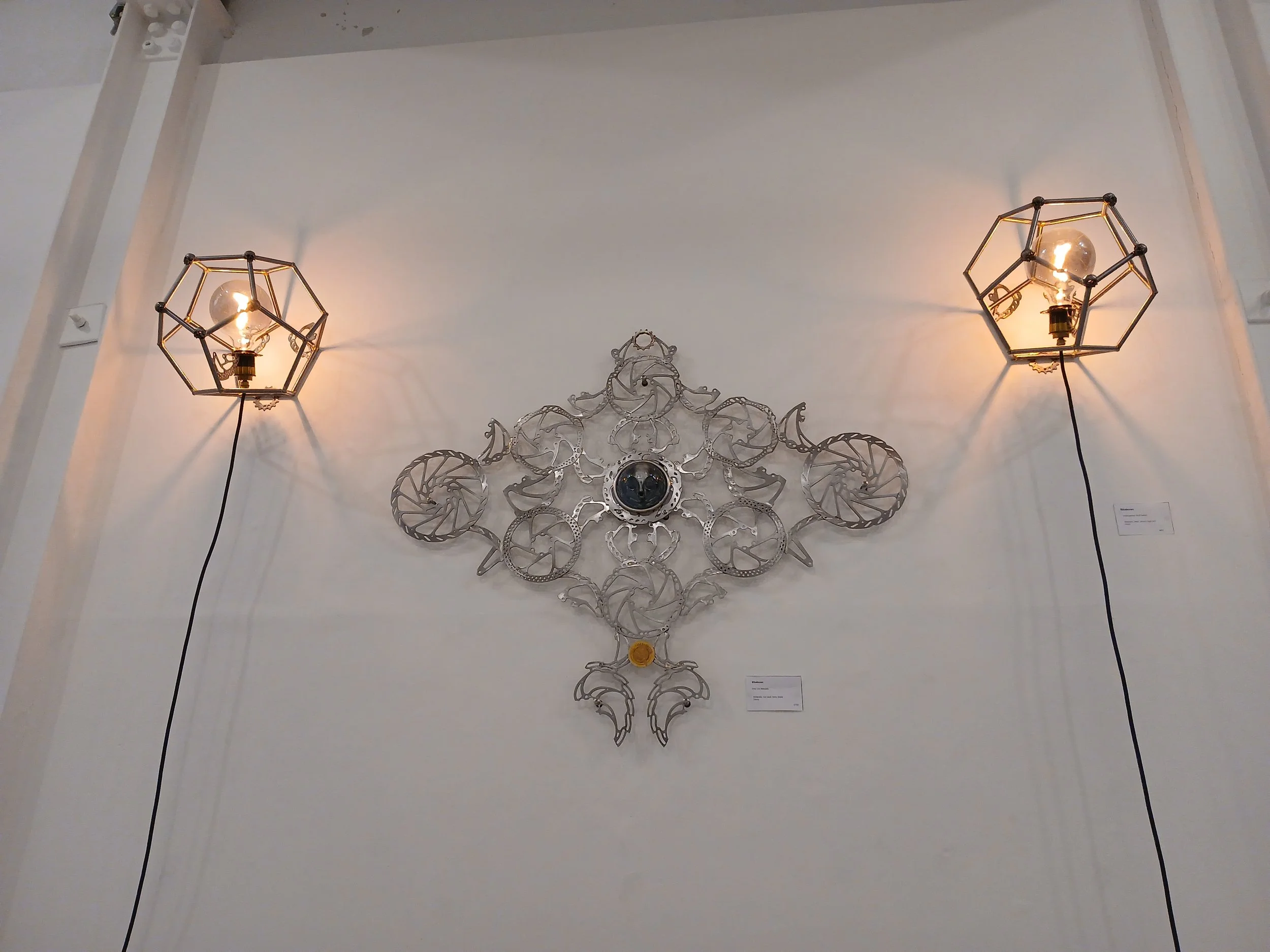 Wall display with two modern hexagonal wall lamps on either side, and a metal decorative art piece in the center, all mounted on a white wall.