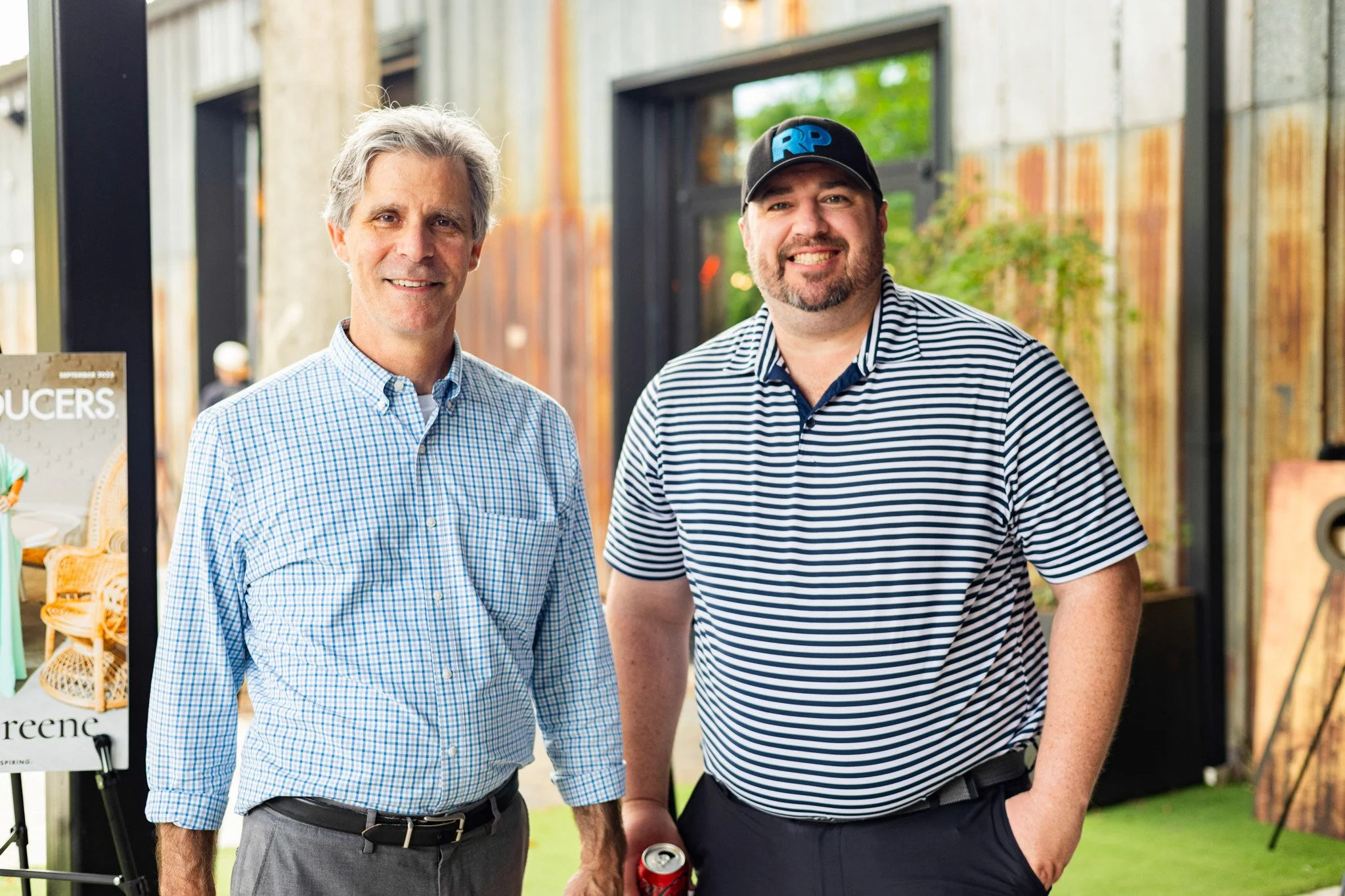 Two men standing outdoors and smiling, one wearing a blue checkered shirt and the other a striped polo shirt with a baseball cap.