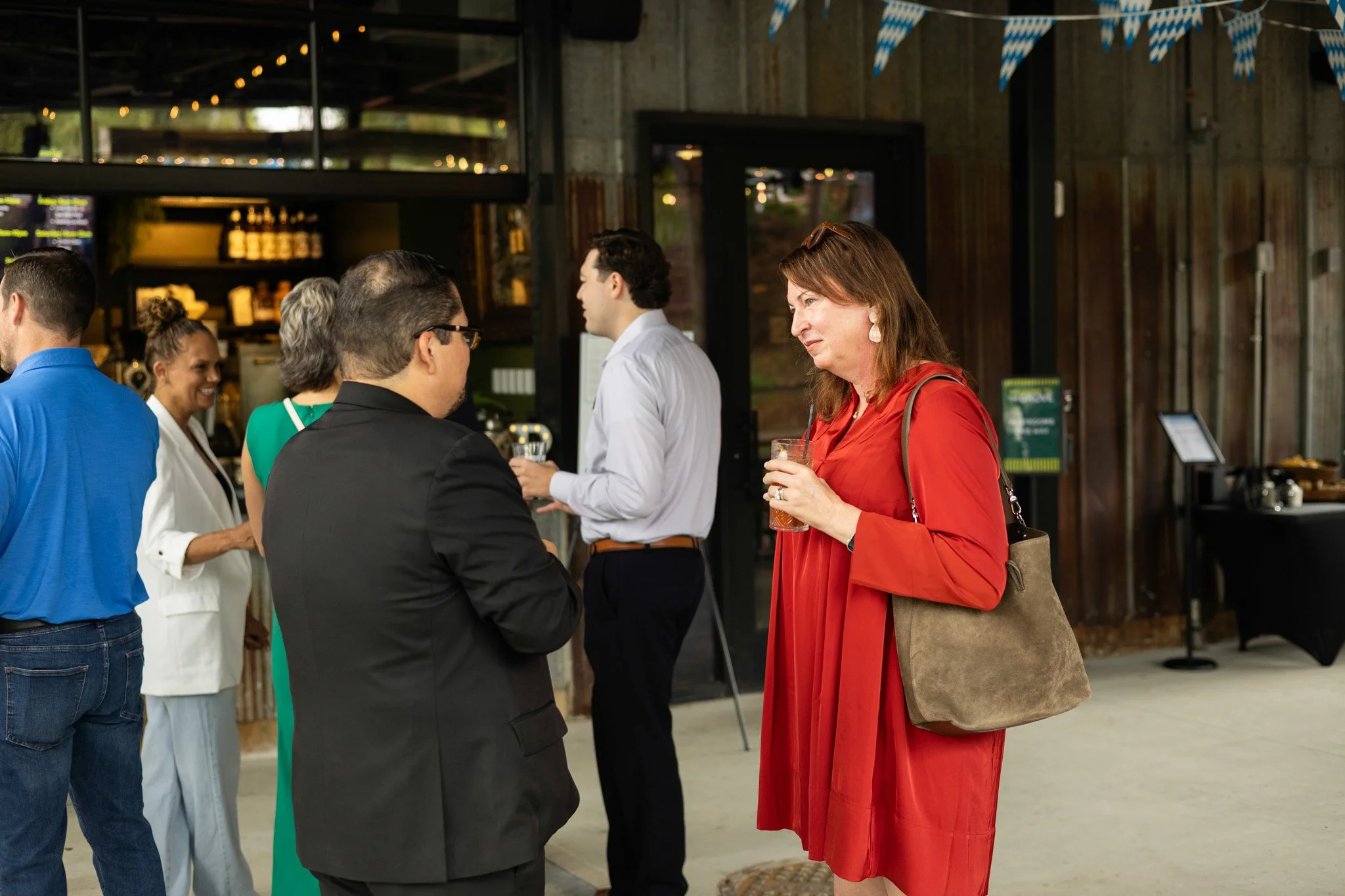 People socializing at an indoor event with drinks, including a woman in a red dress holding a glass and talking to a man in a black suit, with other attendees in the background.