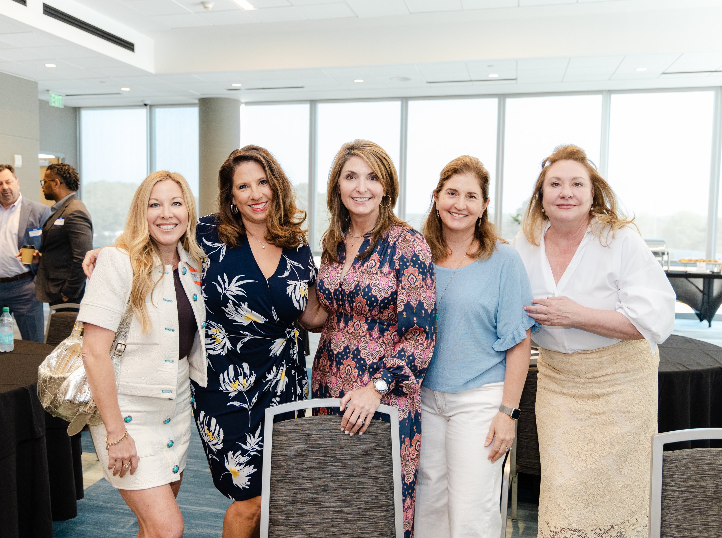 Group of five women smiling and posing together at an indoor event, with large windows in the background.