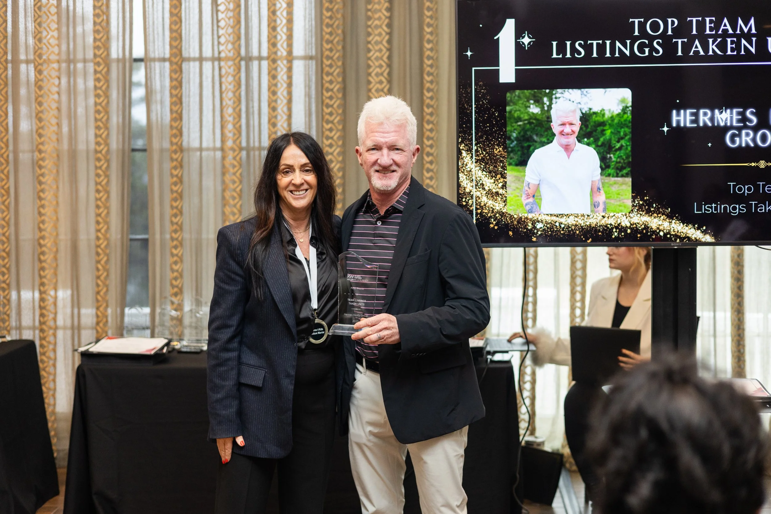 A woman and a man stand together at an award ceremony, smiling at the camera. The woman is wearing a dark suit, and the man is holding a glass award. Behind them, a large screen displays their achievement.