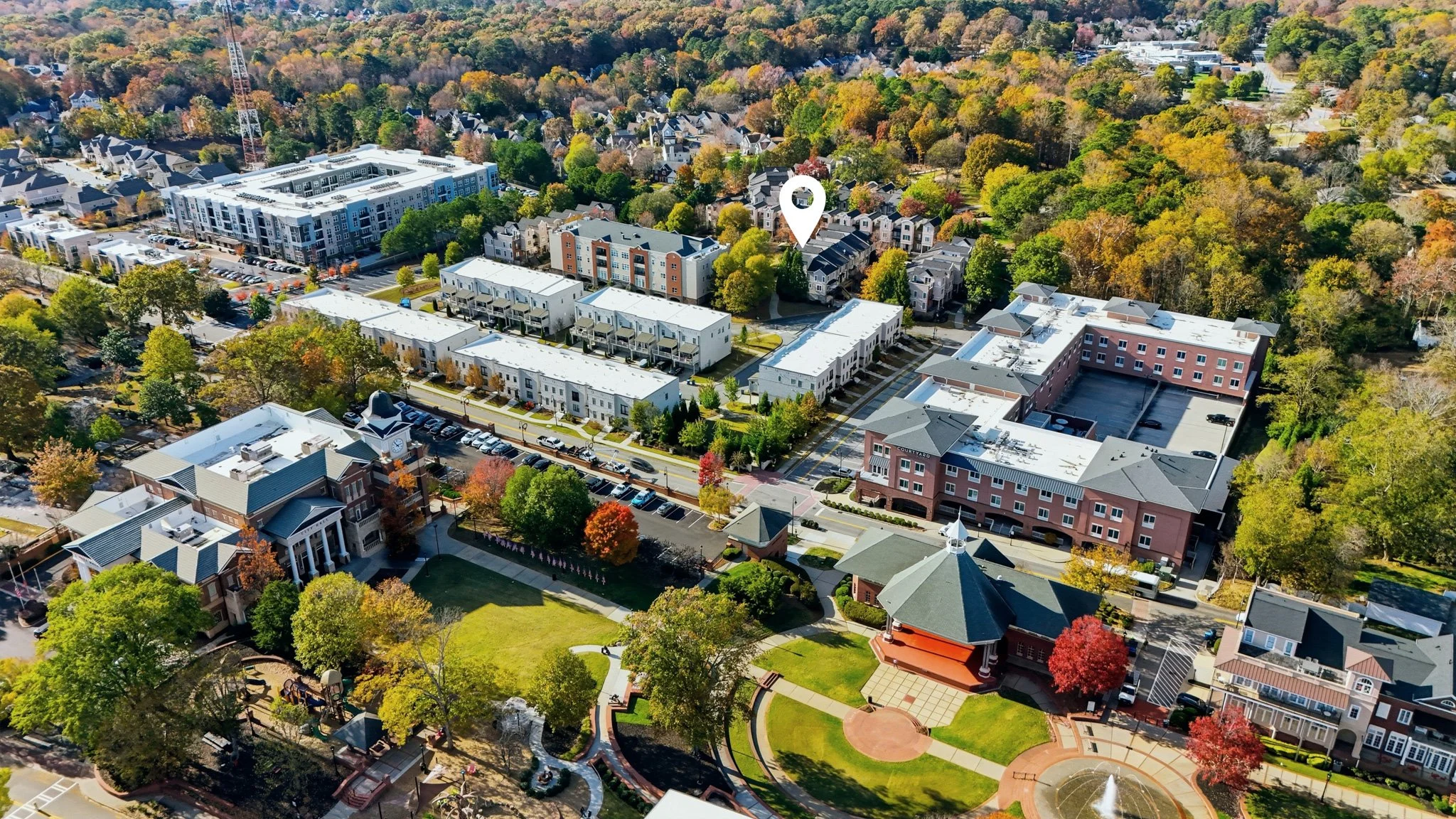 An aerial view of a neighborhood with residential buildings, commercial structures, and a park with a fountain and trees in autumn colors.