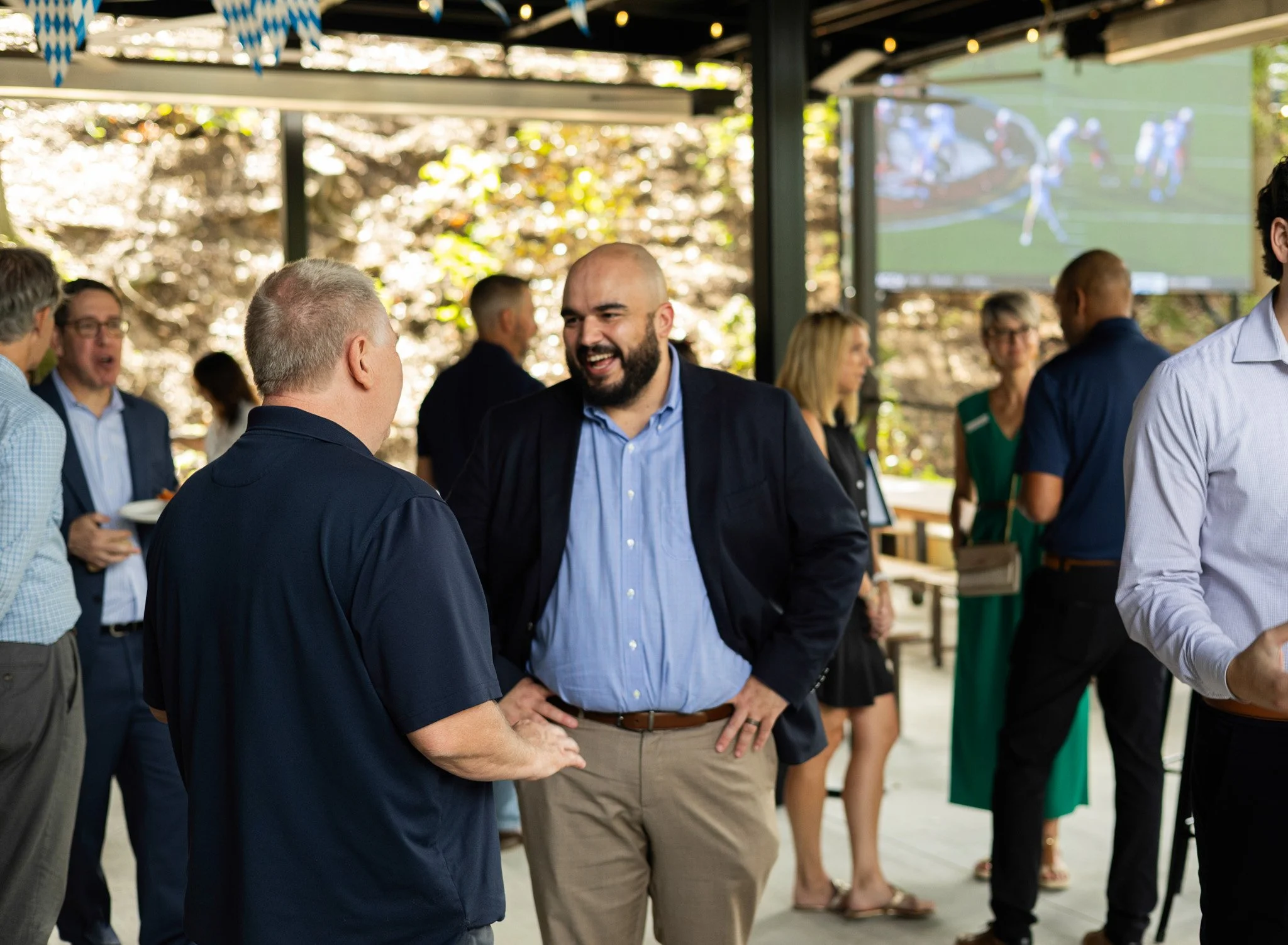 Group of people socializing at an outdoor event with a large screen showing a sports game in the background.