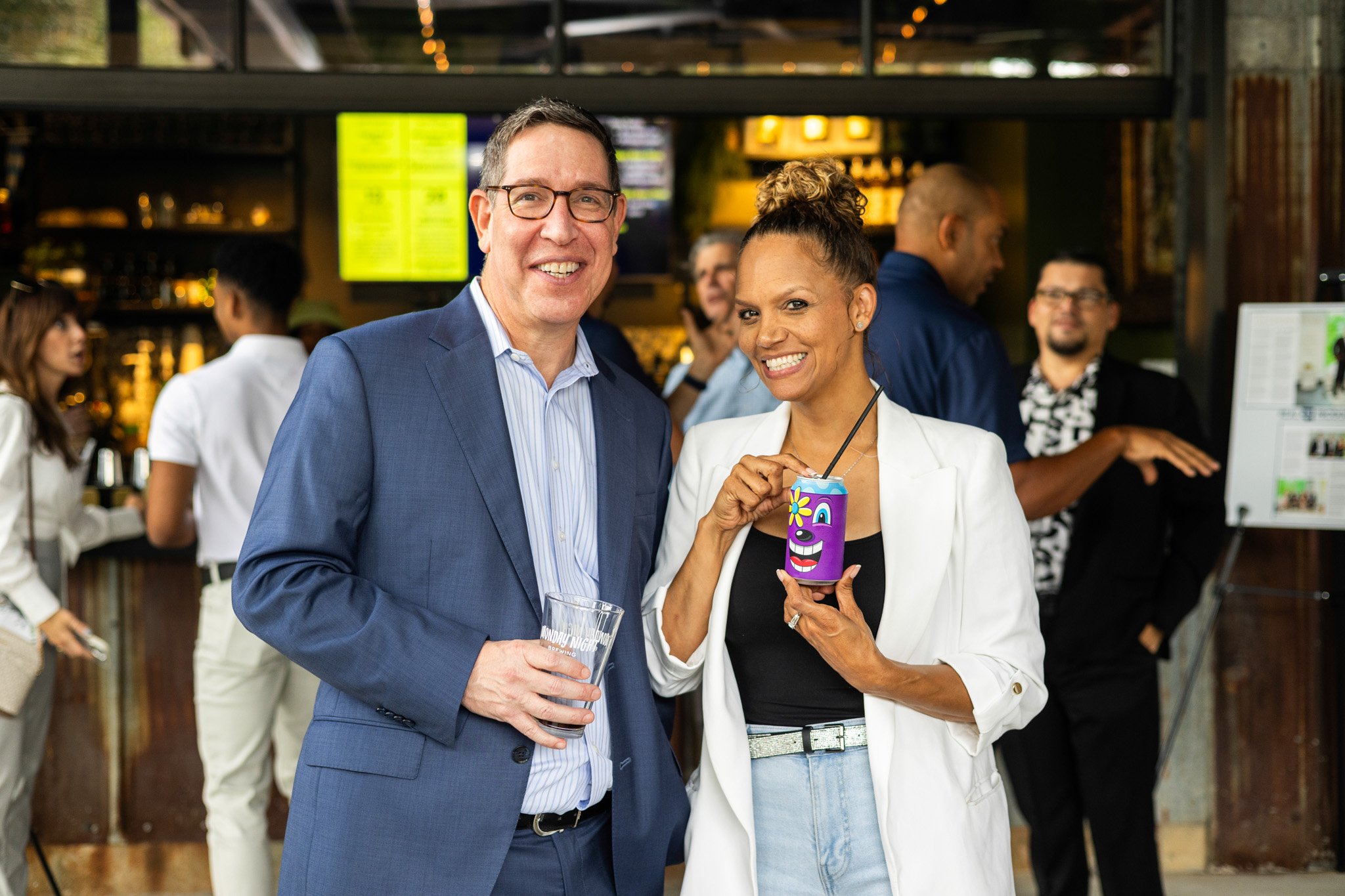 A man in a blue suit and a woman in a white jacket with a black top are smiling and posing for a photo at a social event or gathering in a bar or restaurant. The woman is holding a colorful, cartoon-themed drink.