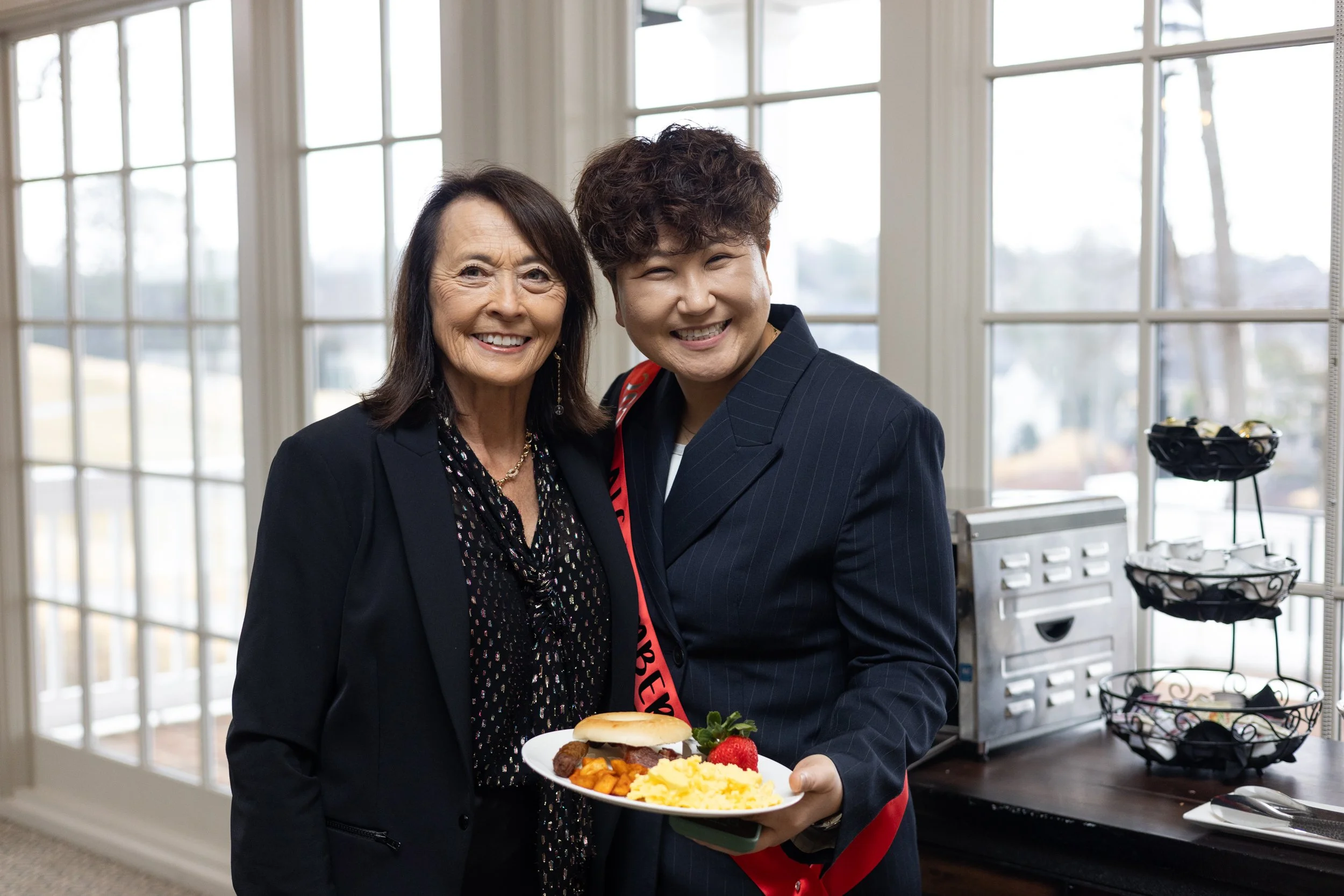 Two women smiling in a room with large windows, one holding a plate of food including scrambled eggs, a hamburger slider, and fruit, the other wearing a sash with red and black writing.