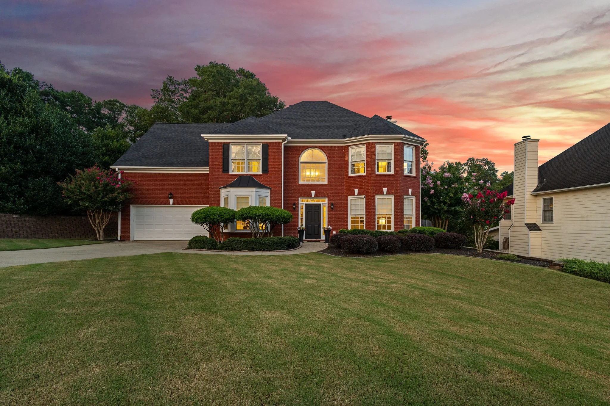 A large two-story brick house with multiple windows and a black front door, surrounded by a well-maintained lawn and shrubs, with a sunset sky in the background.