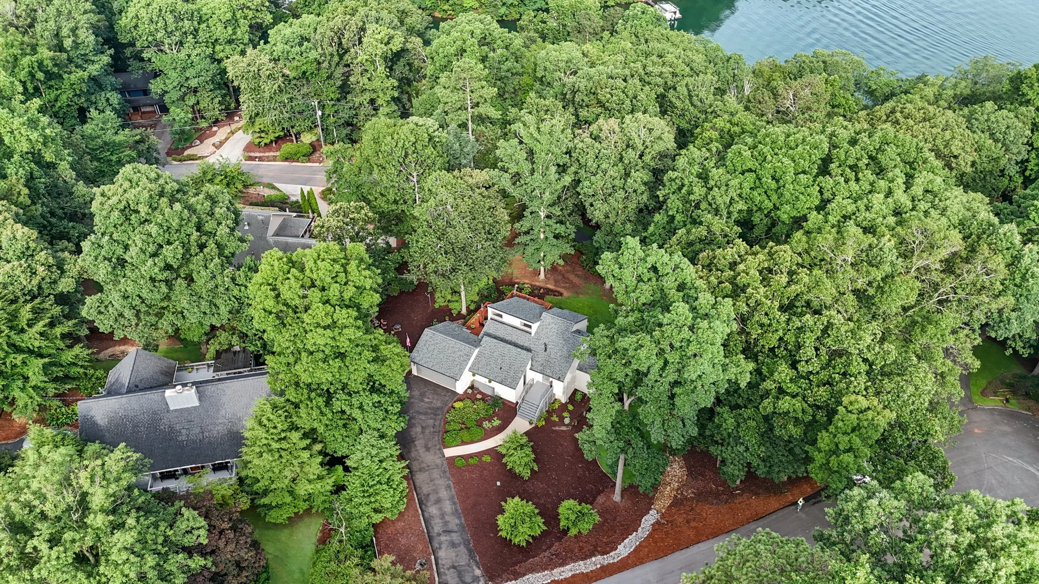 An aerial view of a residential neighborhood surrounded by dense green trees, with houses, driveways, and a nearby body of water in the background.