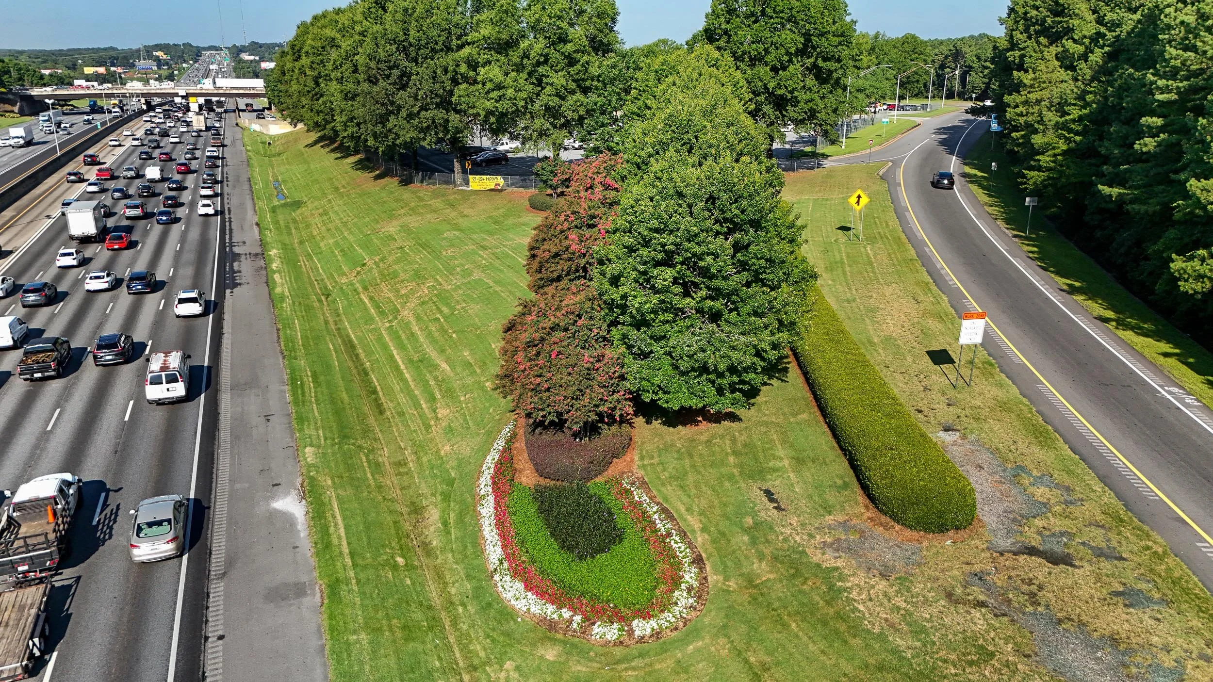 An overhead view of a highway with flowing traffic on the left, and a empty curving road on the right separated by a green grass median with trees and a flower bed.