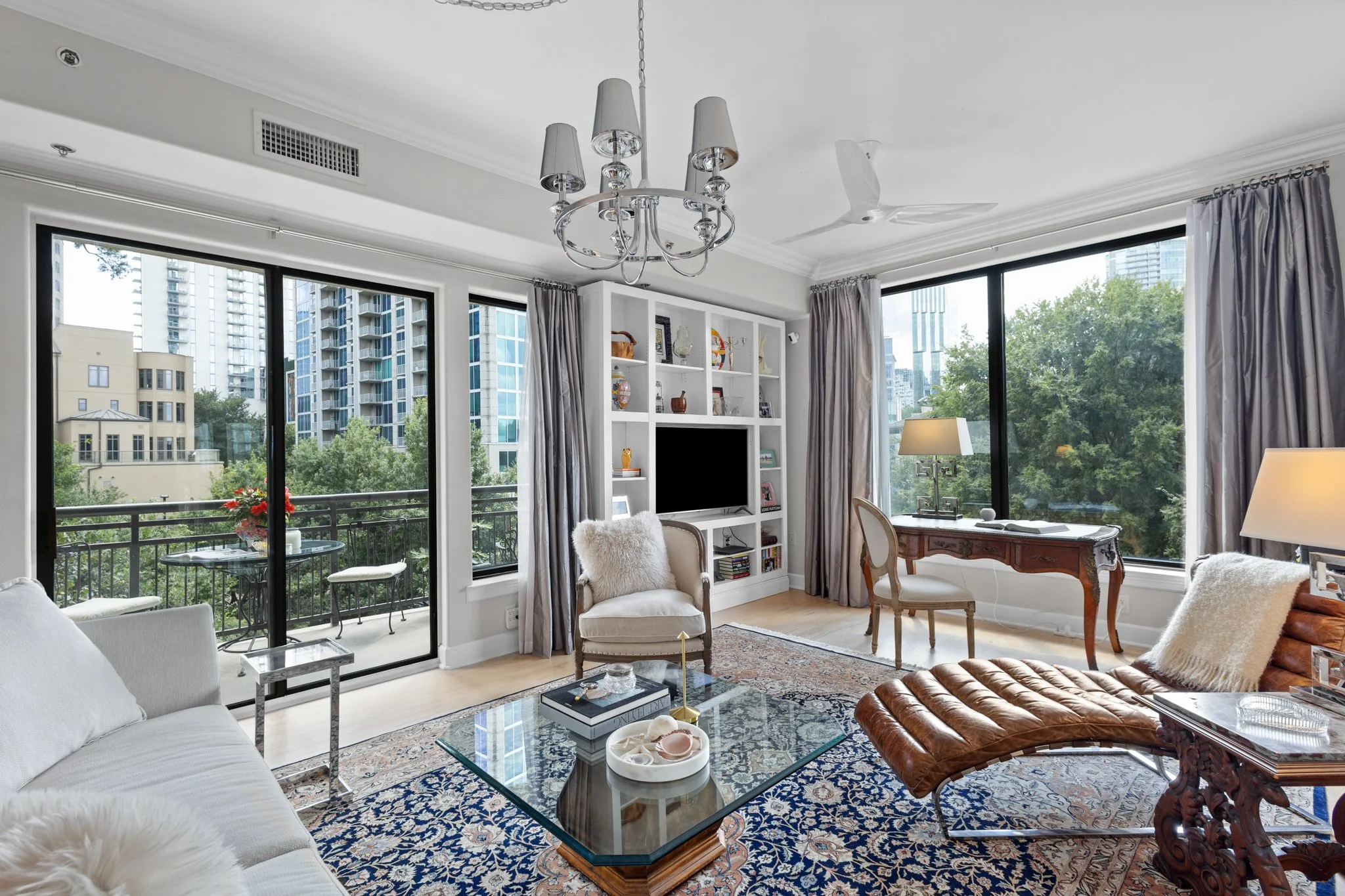 Living room with large windows, a glass coffee table, white and beige furniture, a wooden writing desk, and a balcony with outdoor seating. Decor includes a chandelier, lamps, and decorative items on white shelving.