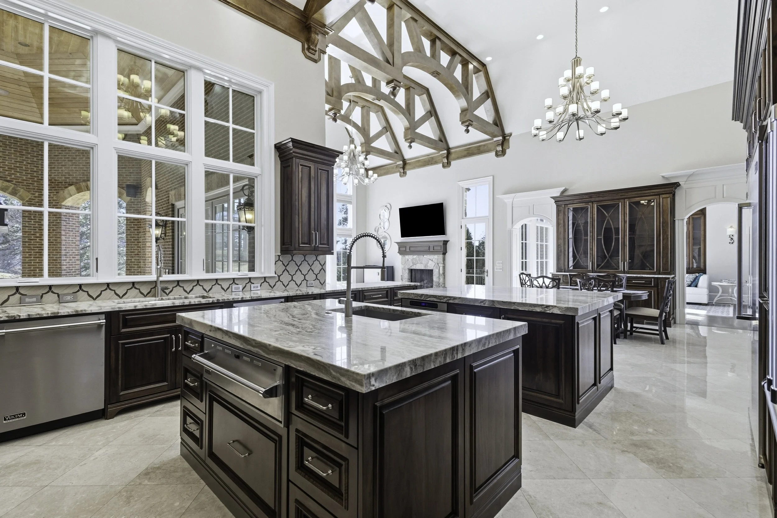 Modern kitchen with dark wooden cabinets, an island with marble countertop, large windows, a chandelier, a fireplace, and a flat-screen TV above it.