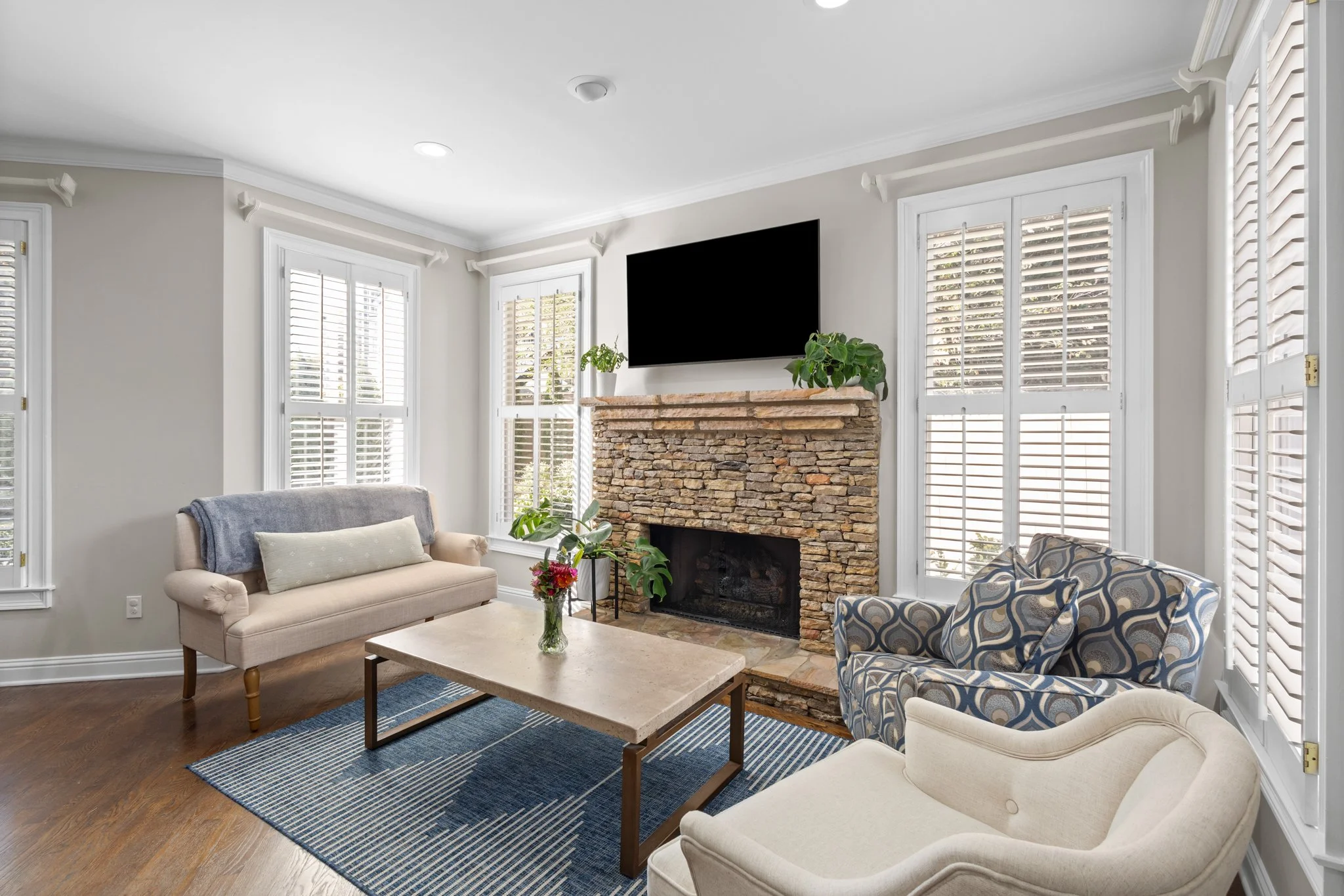 Living room with beige, patterned, and blue armchairs, a coffee table, a fireplace with a stone mantel, a flat-screen TV above the fireplace, and three large windows with white blinds.