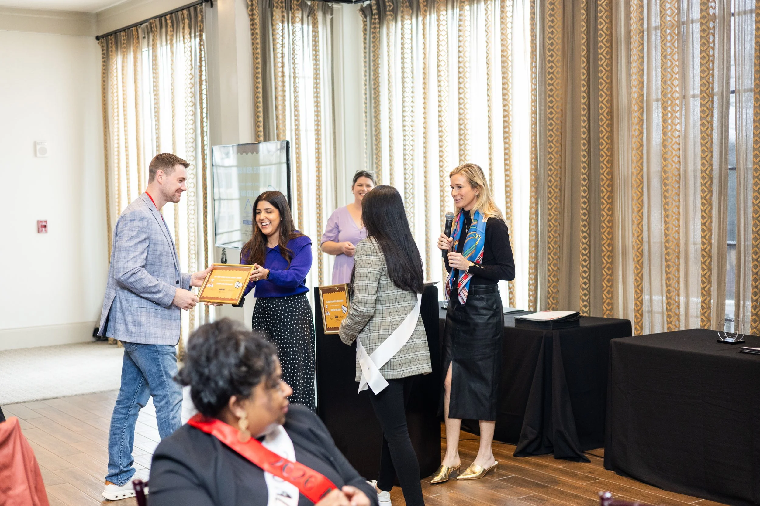A group of people in a conference room during an award ceremony. A man in a gray blazer receives a certificate from a woman in a blue dress. Two women, one with long black hair in a gray plaid jacket and one with blonde hair holding a microphone, are