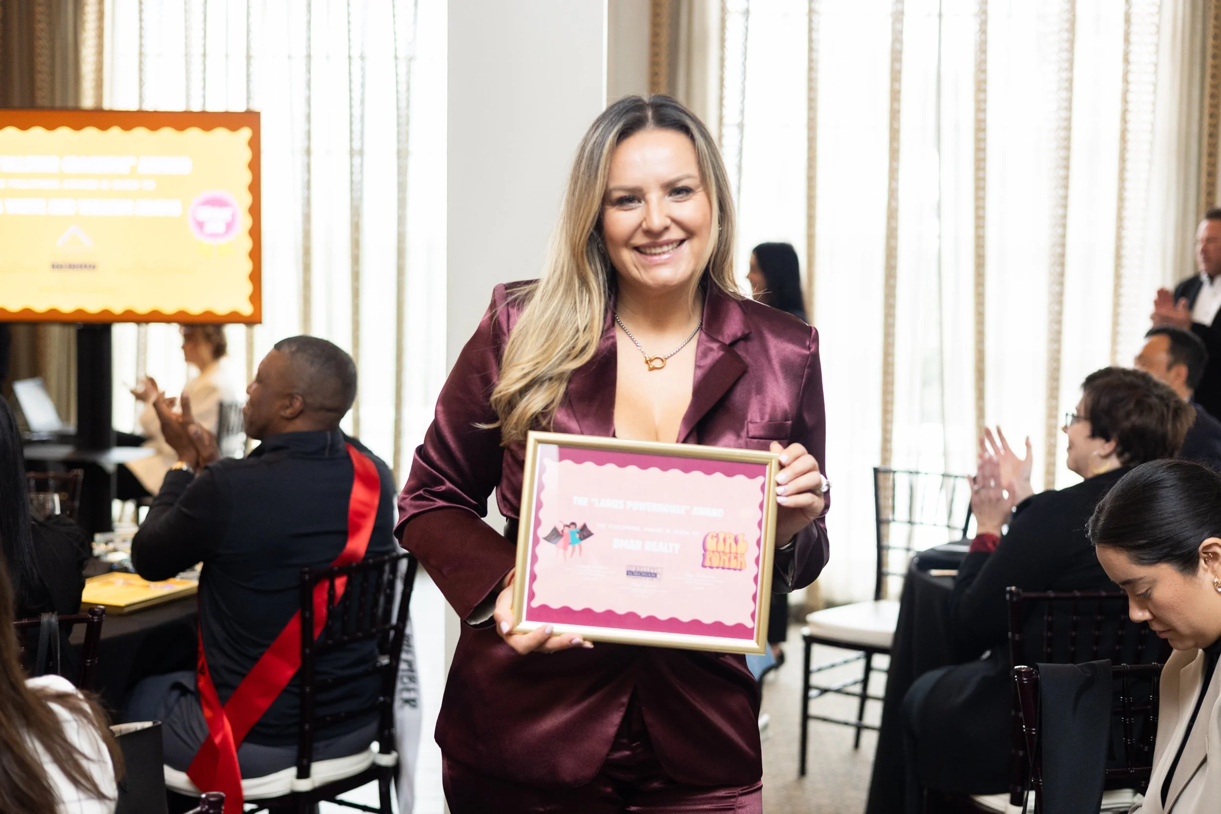 A woman in maroon suit holding a framed award, smiling at a formal event with people sitting around, some clapping, in a bright room with large curtains.