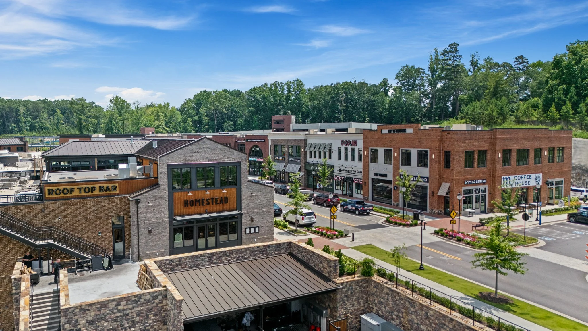 An aerial view of a small shopping and dining area with brick and modern buildings, parking lot with cars, trees, and a background of lush green forest and blue sky.