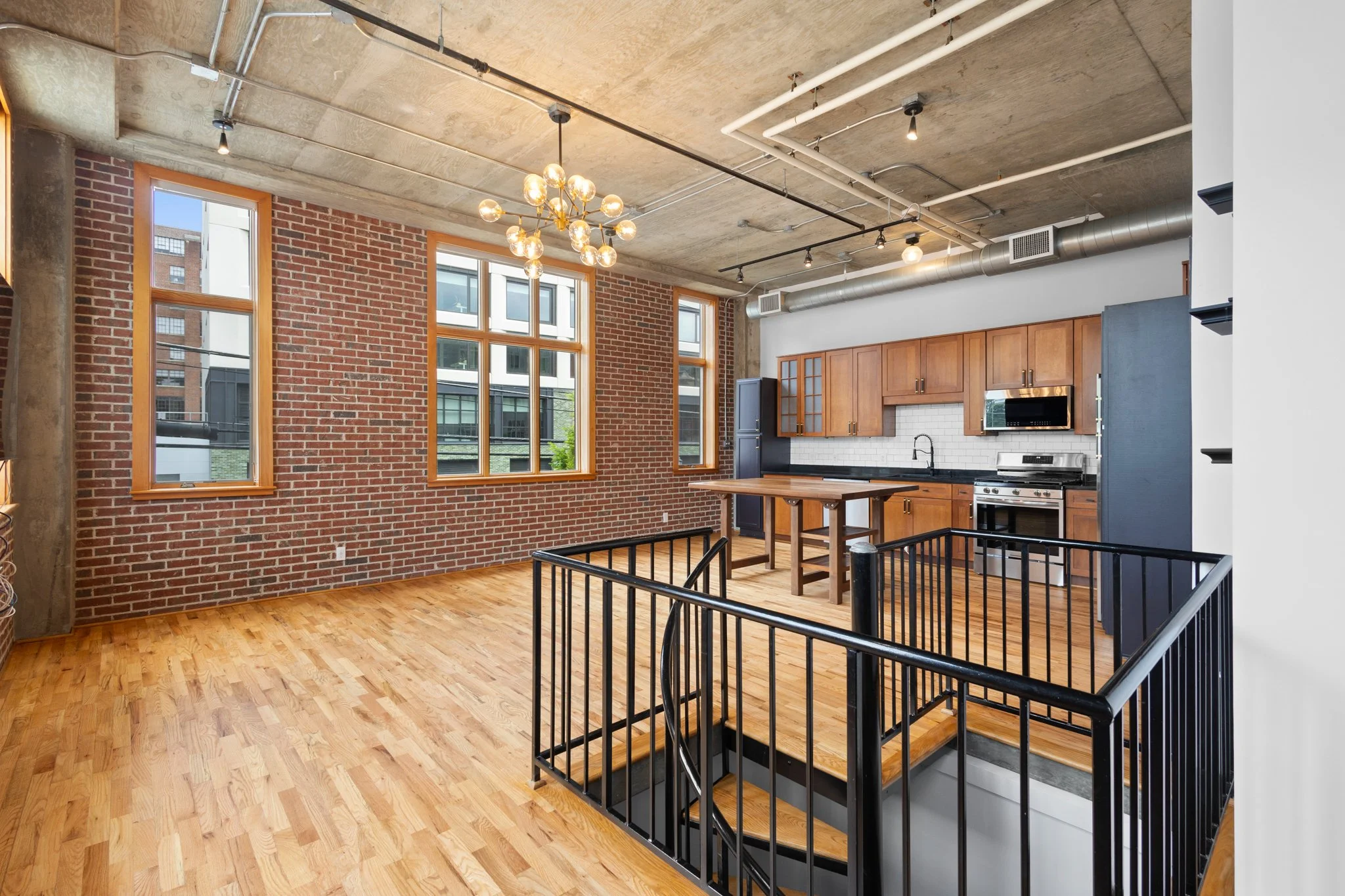 Open-concept kitchen and living space with brick walls, wood flooring, large windows, modern ceiling lighting, and an industrial-style ceiling.