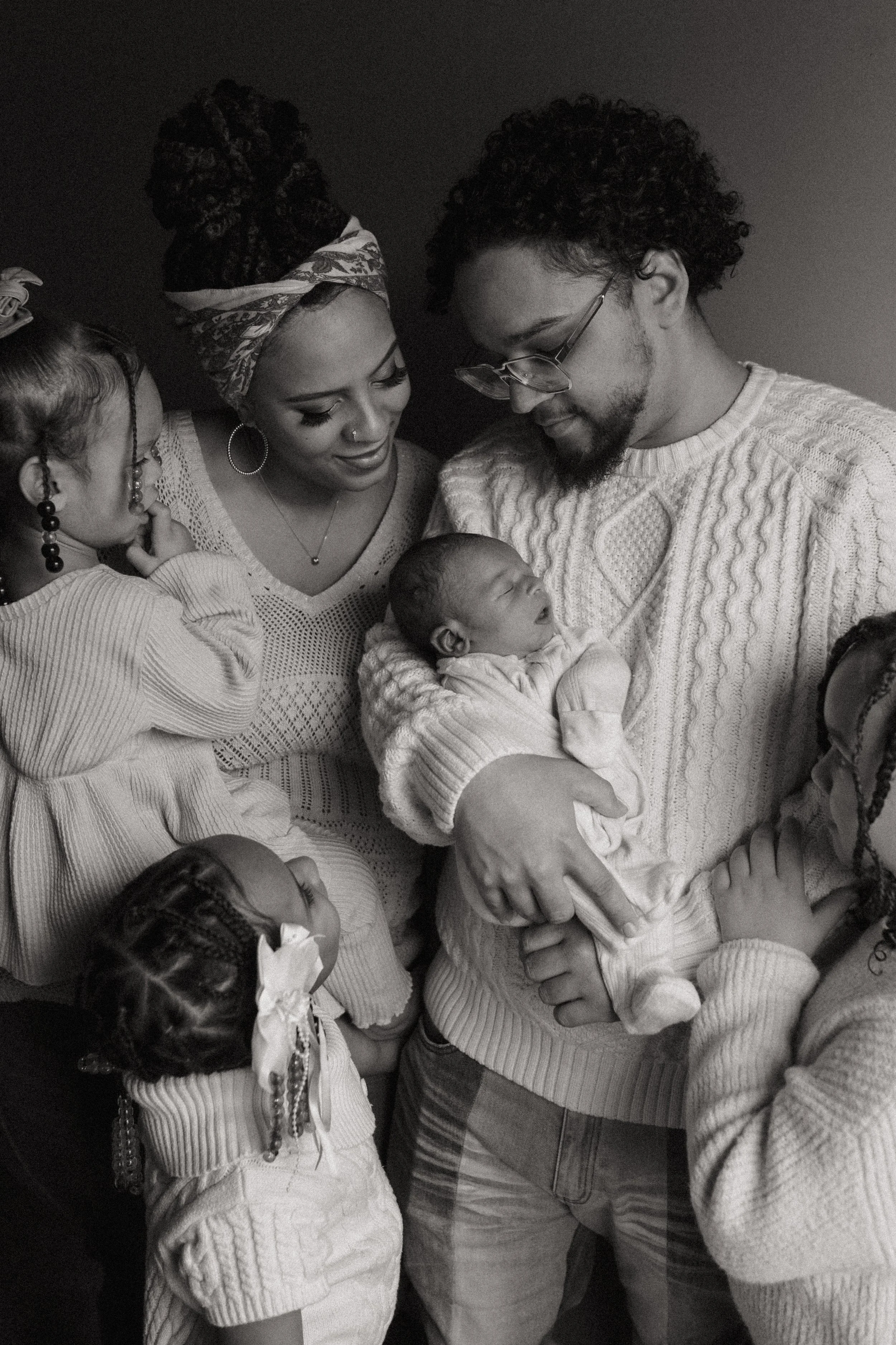 A black and white photo of a diverse family holding a newborn baby, with children gathered around. The adults look at the baby lovingly, and the children look on with interest.