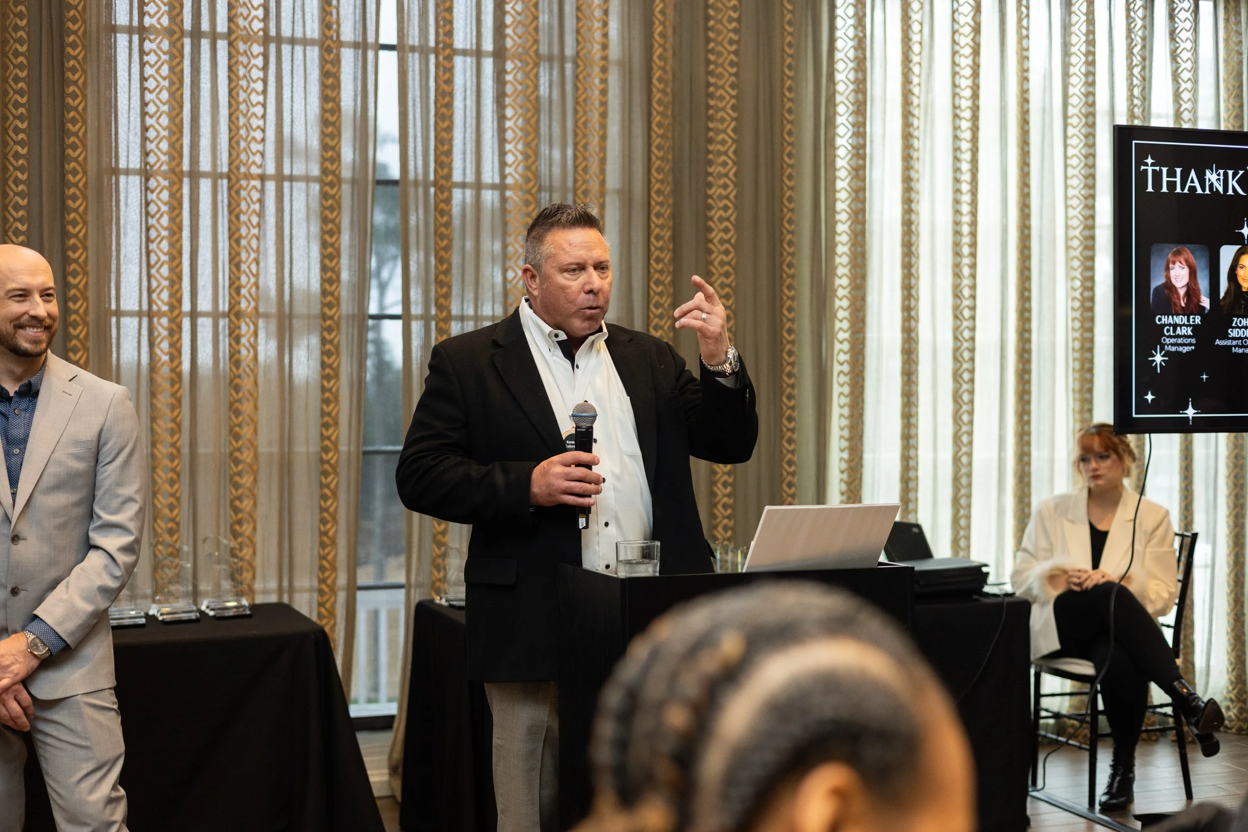 Man in black blazer speaking into microphone at a conference, with a laptop on the podium, a woman sitting to the right and a large screen displaying a thank you message with names and photos of individuals in the background.