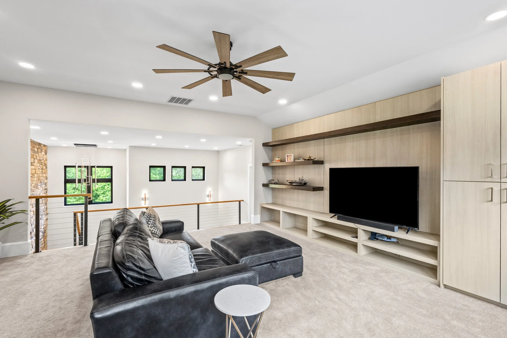 Living room with black leather sofa, beige carpet, wall-mounted TV, light wood entertainment unit, floating shelves, ceiling fan, and large windows with greenery outside.